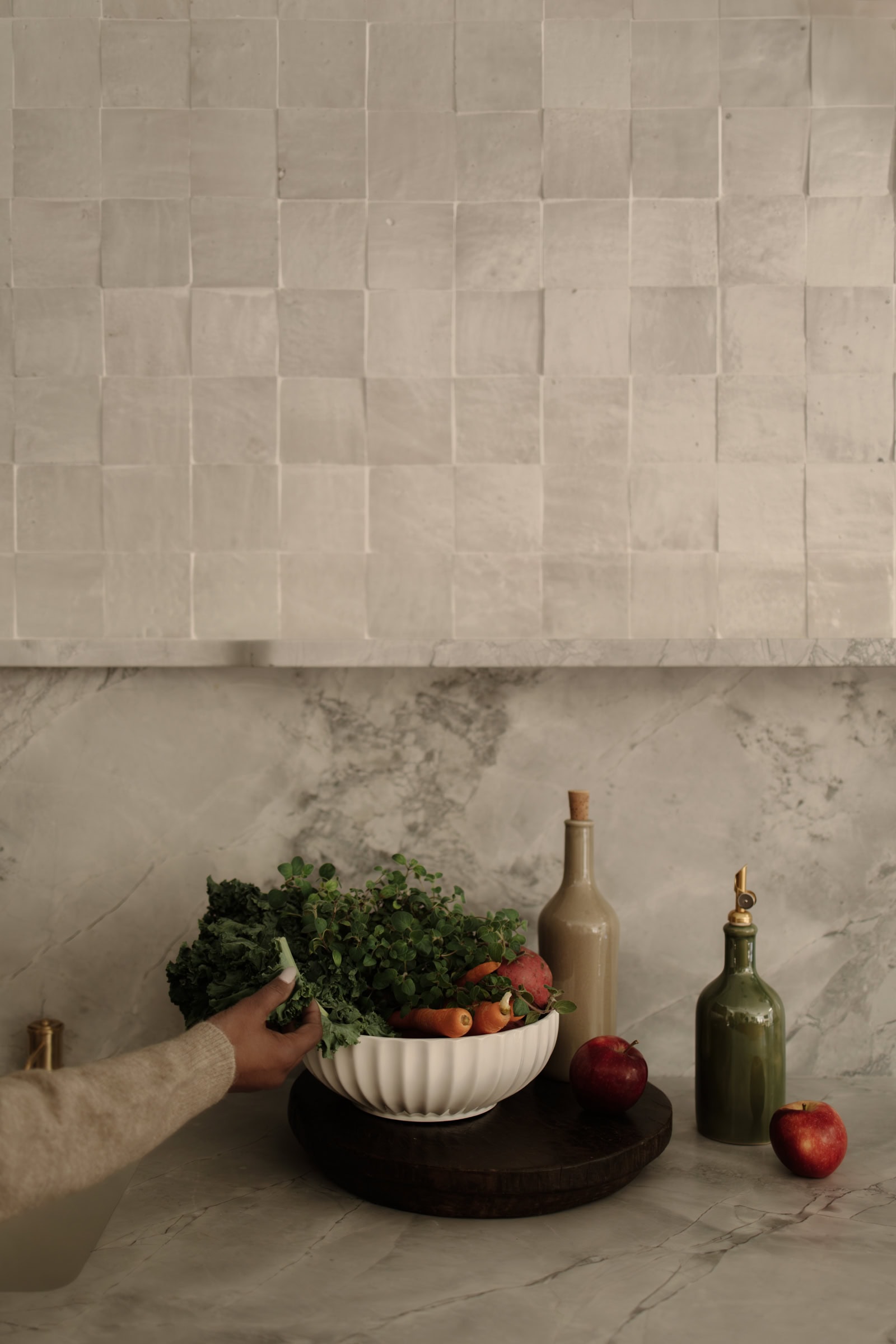 A hand reaches for leafy greens in a white bowl filled with vegetables and fruits on a marble countertop, next to two glass bottles and two apples, with a textured light-colored tile backsplash.