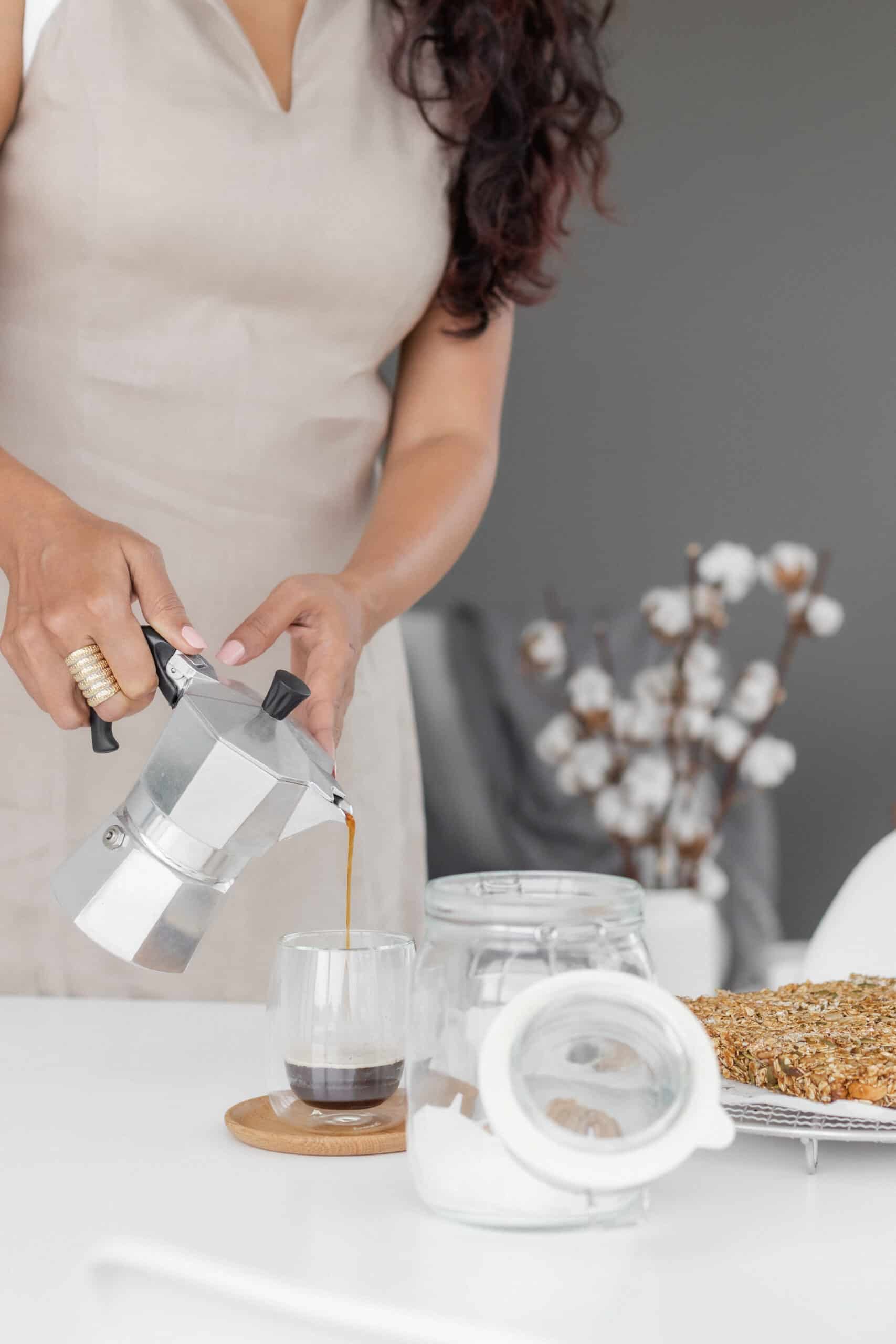 A woman in a beige dress pours coffee from a moka pot into a glass on a white table, embodying the snug and cozy life. An open jar and a tray of snacks sit nearby, while a blurred cotton plant completes the tranquil scene.