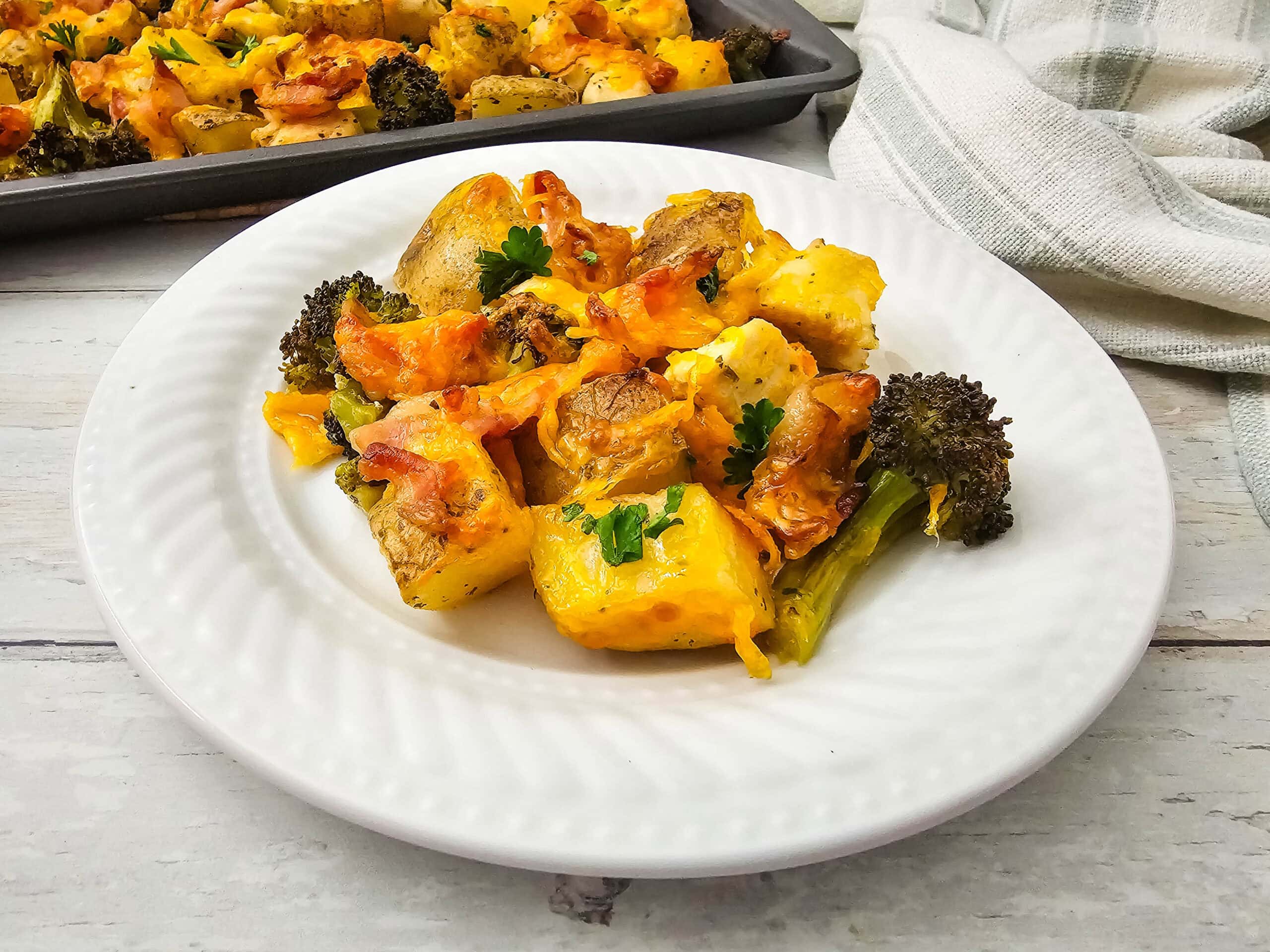 A white plate with roasted potatoes, broccoli, and melted cheese, garnished with parsley. A baking tray with more of the dish and a light cloth are in the background.