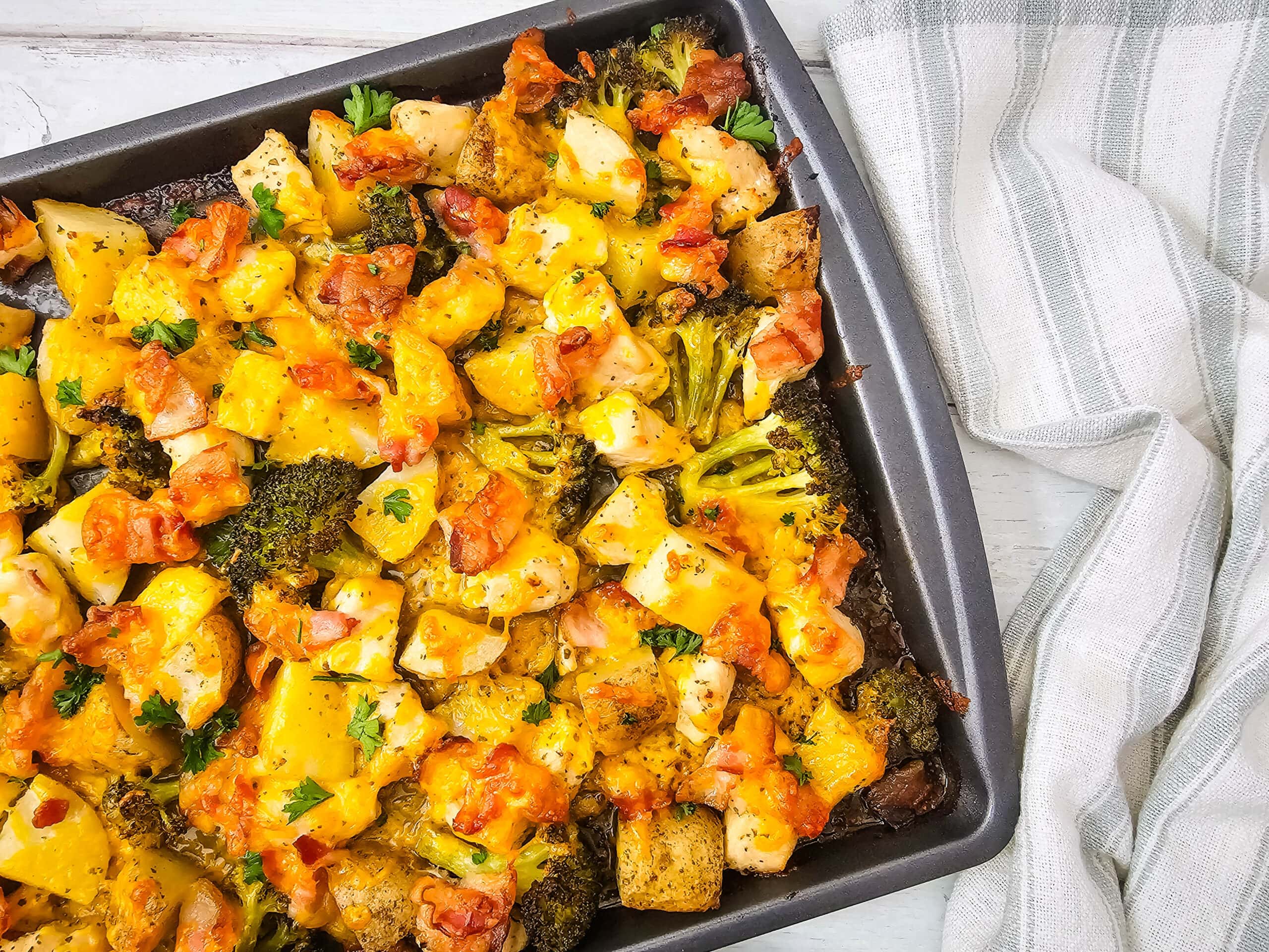 A baking pan filled with a colorful baked dish featuring chunks of chicken, broccoli, potatoes, melted cheese, and bits of tomato, garnished with herbs, sits on a white surface next to a striped kitchen towel.