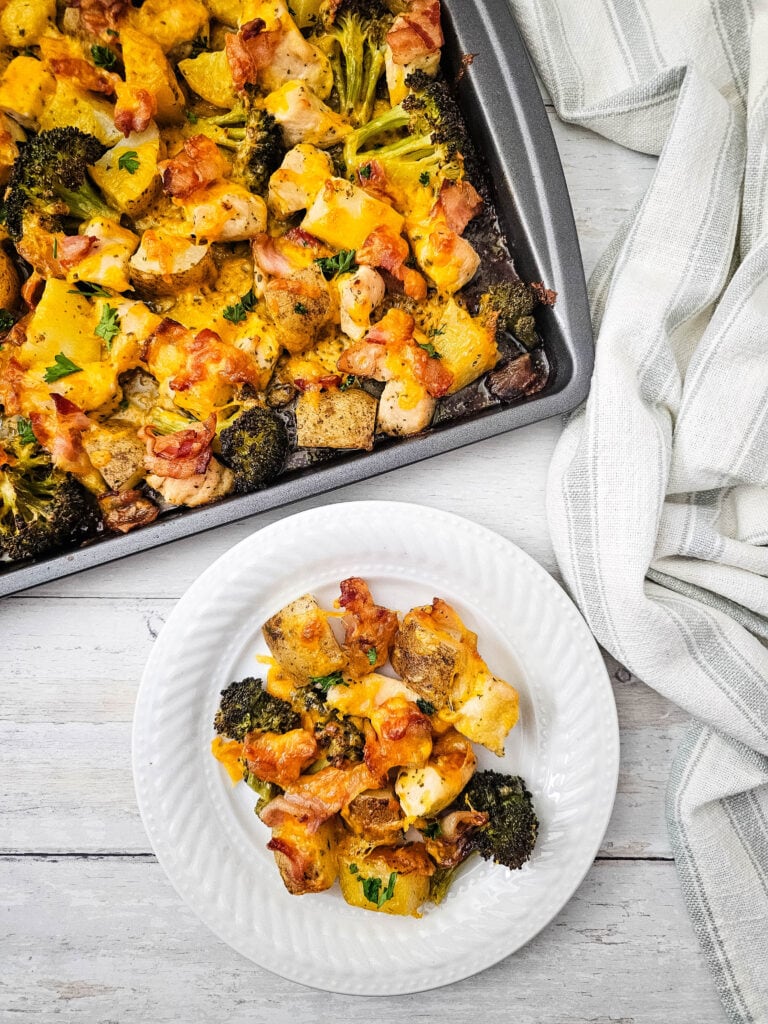 A white plate with a serving of baked chicken, potatoes, broccoli, bacon, and melted cheese sits next to a baking tray with the same dish on a light wooden surface, with a striped kitchen towel nearby.