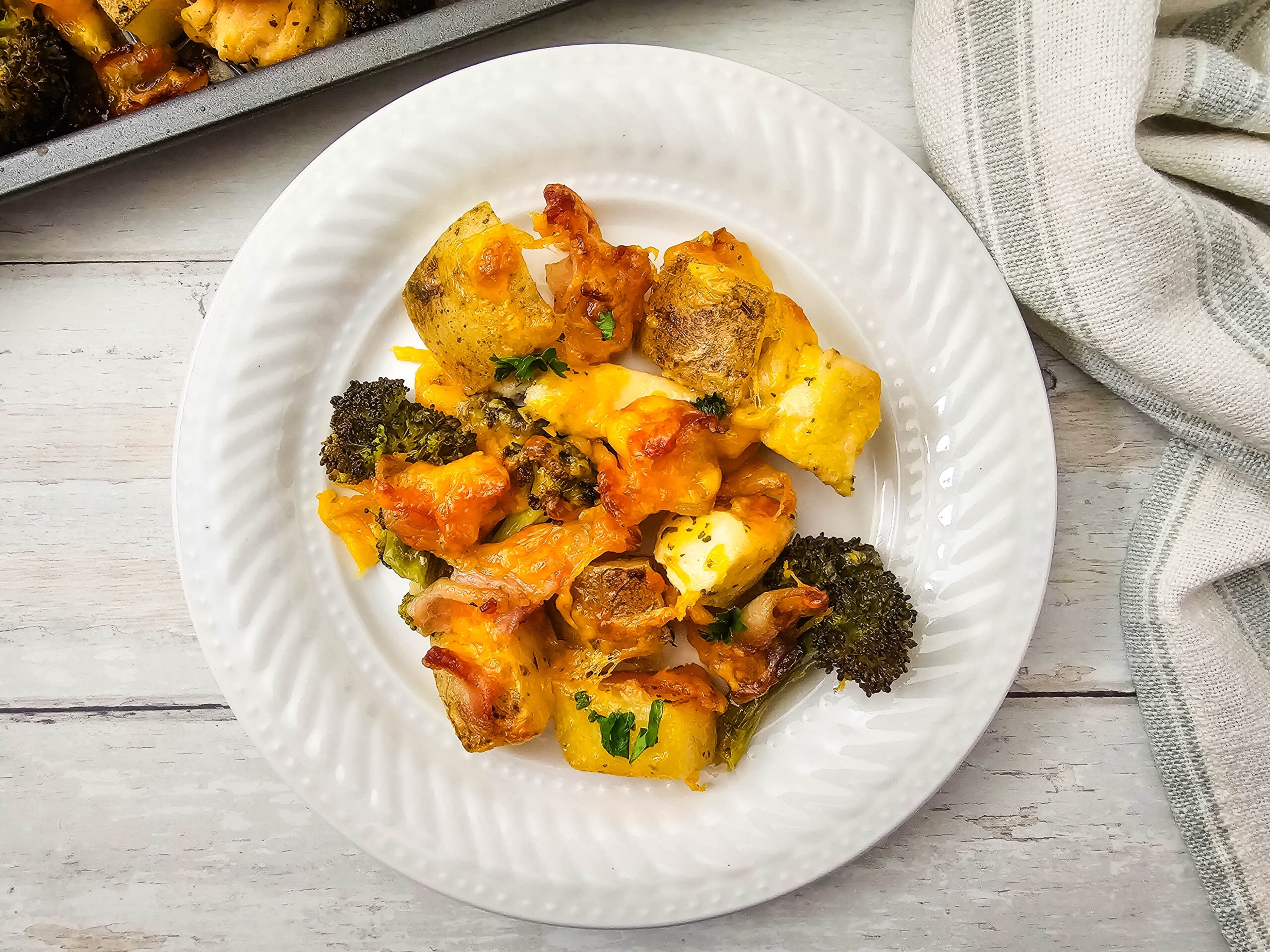 A white plate with baked chicken, potatoes, broccoli, and melted cheese, garnished with herbs, sits on a light wooden table next to a beige-striped cloth.