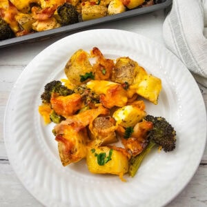 A white plate with a serving of roasted potatoes, broccoli, and chicken pieces topped with melted cheese, garnished with parsley. A baking tray with more of the dish is visible in the background.