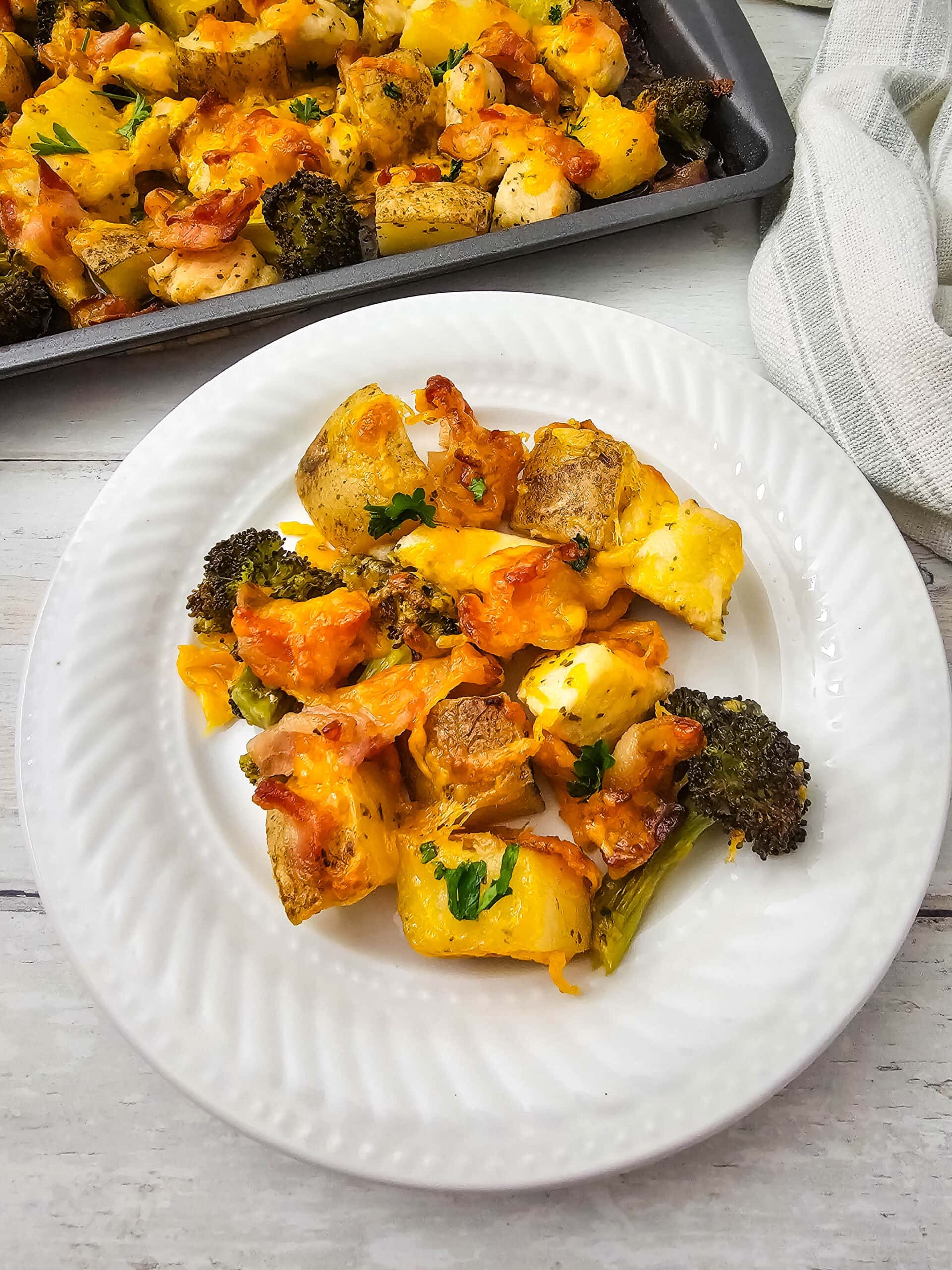 A white plate with a serving of roasted potatoes, broccoli, and chicken pieces topped with melted cheese, garnished with parsley. A baking tray with more of the dish is visible in the background.