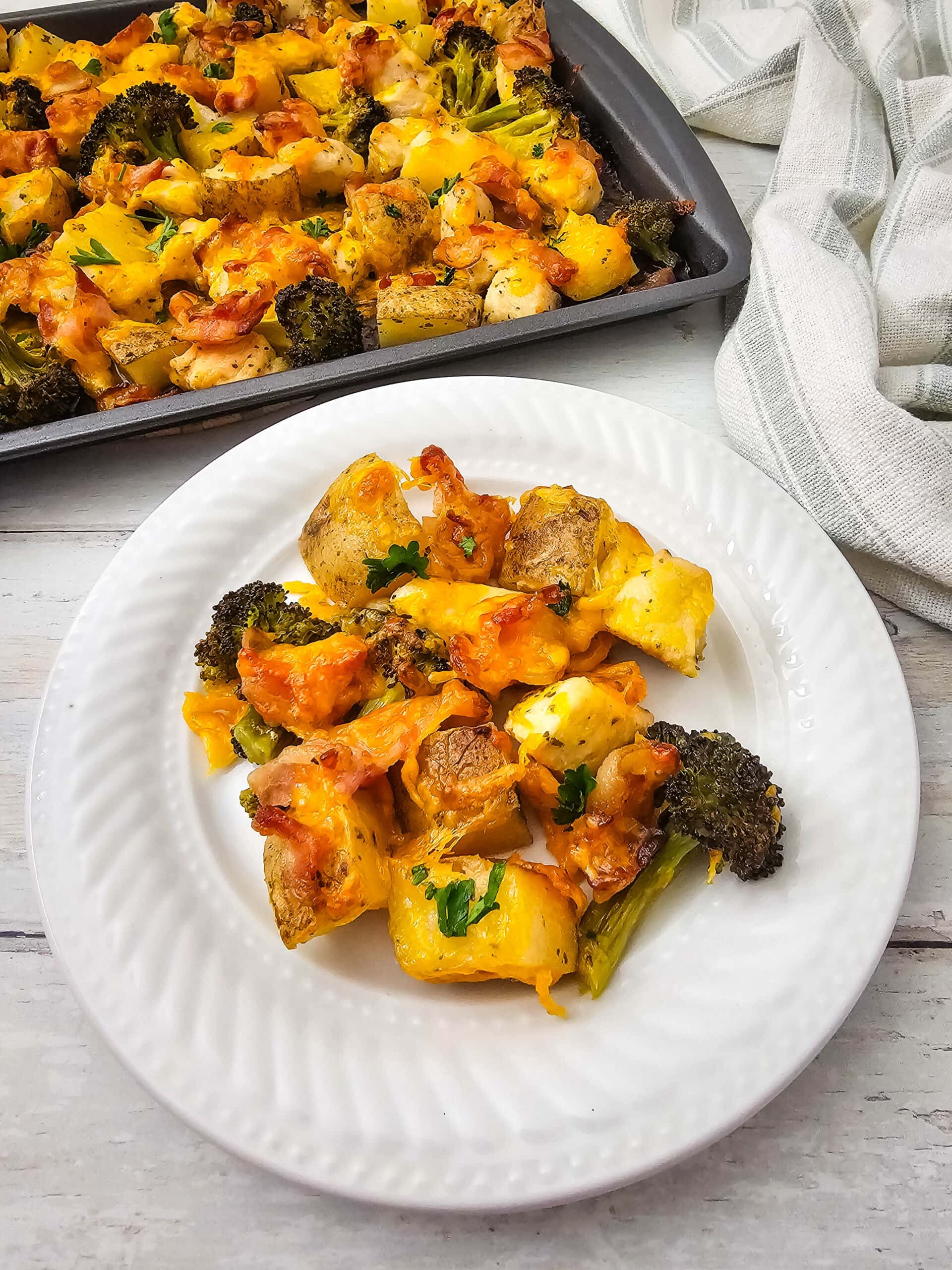 A white plate with a serving of baked chicken, potatoes, broccoli, and melted cheese, garnished with herbs. In the background, a baking tray with more of the same dish sits on a light wooden surface.