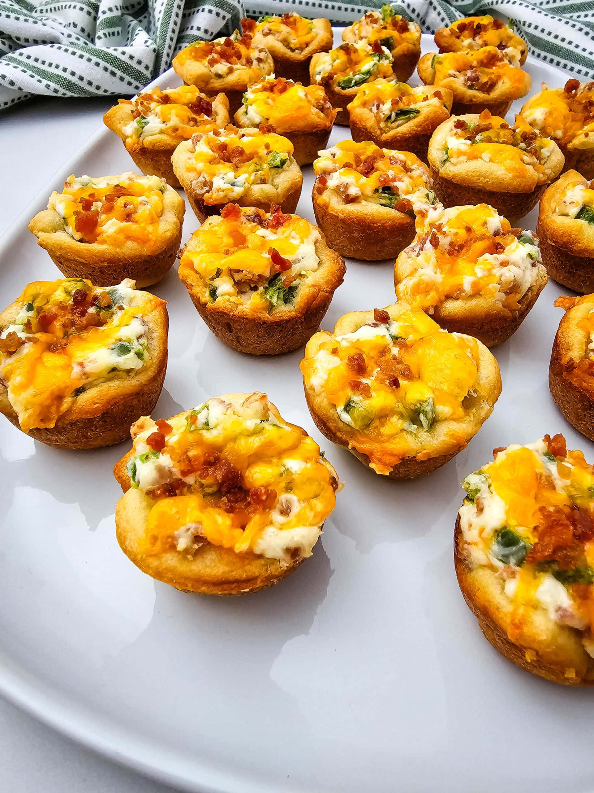 A white plate filled with bite-sized baked appetizers topped with melted cheddar cheese, bacon bits, and green onions. A green and white patterned cloth is visible in the background.