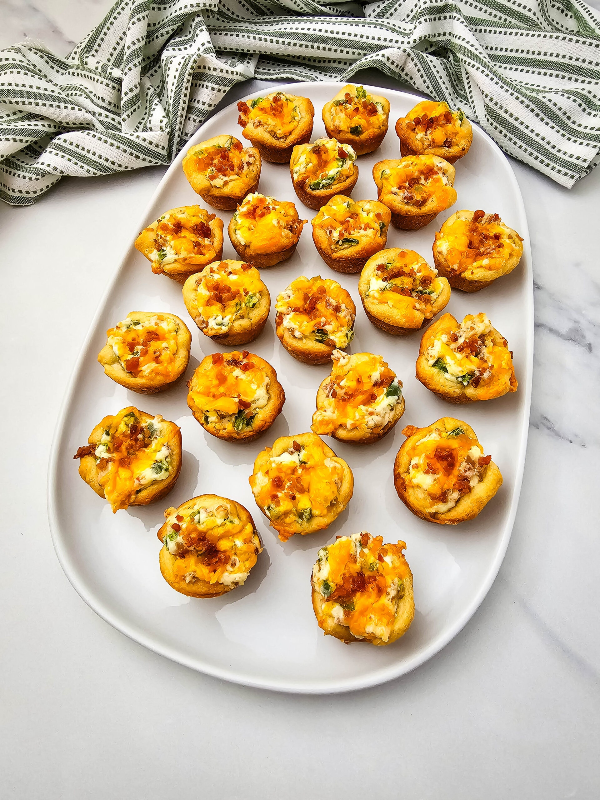 A white oval platter holds 21 mini savory muffin cups topped with melted cheese and bits of vegetables. A green-and-white striped cloth is in the background on a marble surface.