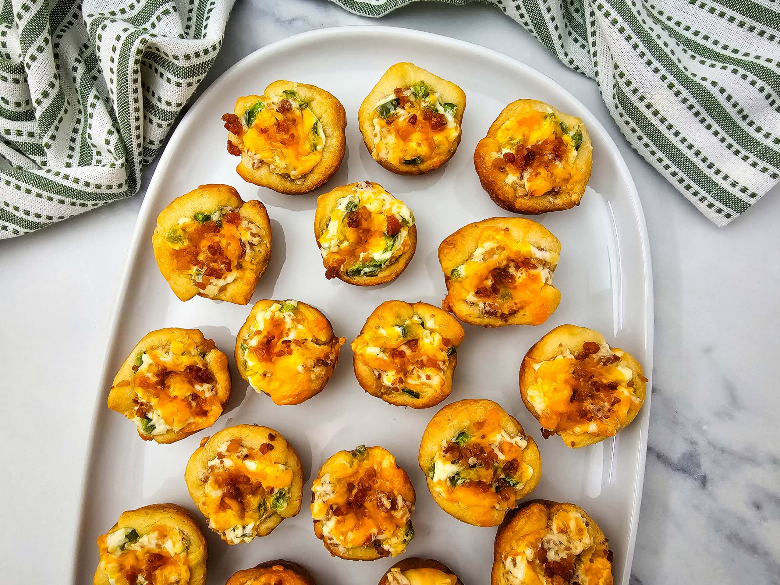 A white plate topped with several mini biscuit cups filled with cheese, bacon, and green onions. A green and white striped cloth napkin is partially visible in the background on a marble surface.