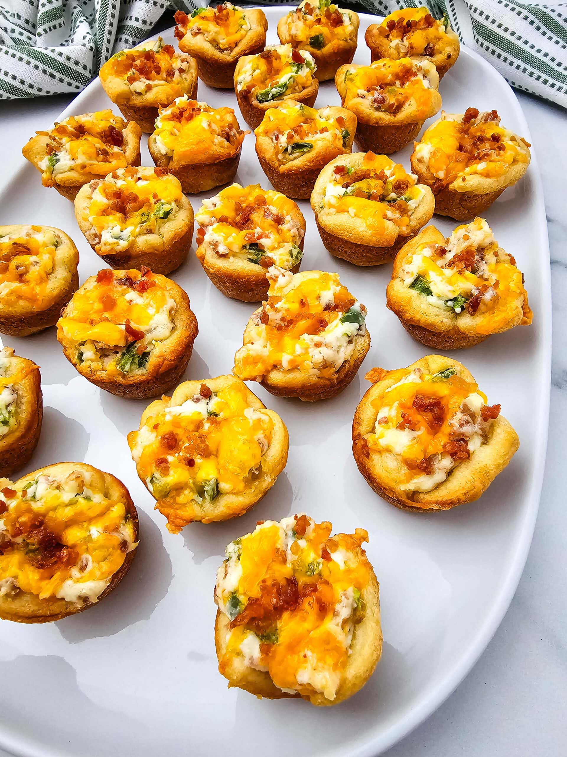 A white platter holds several bite-sized savory muffins topped with melted cheese, bacon bits, and chopped green herbs. The background features part of a striped cloth.