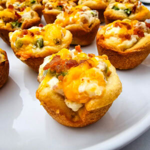 A close-up of savory baked muffin cups filled with creamy cheese, chopped green onions, and bacon bits, arranged on a white plate.