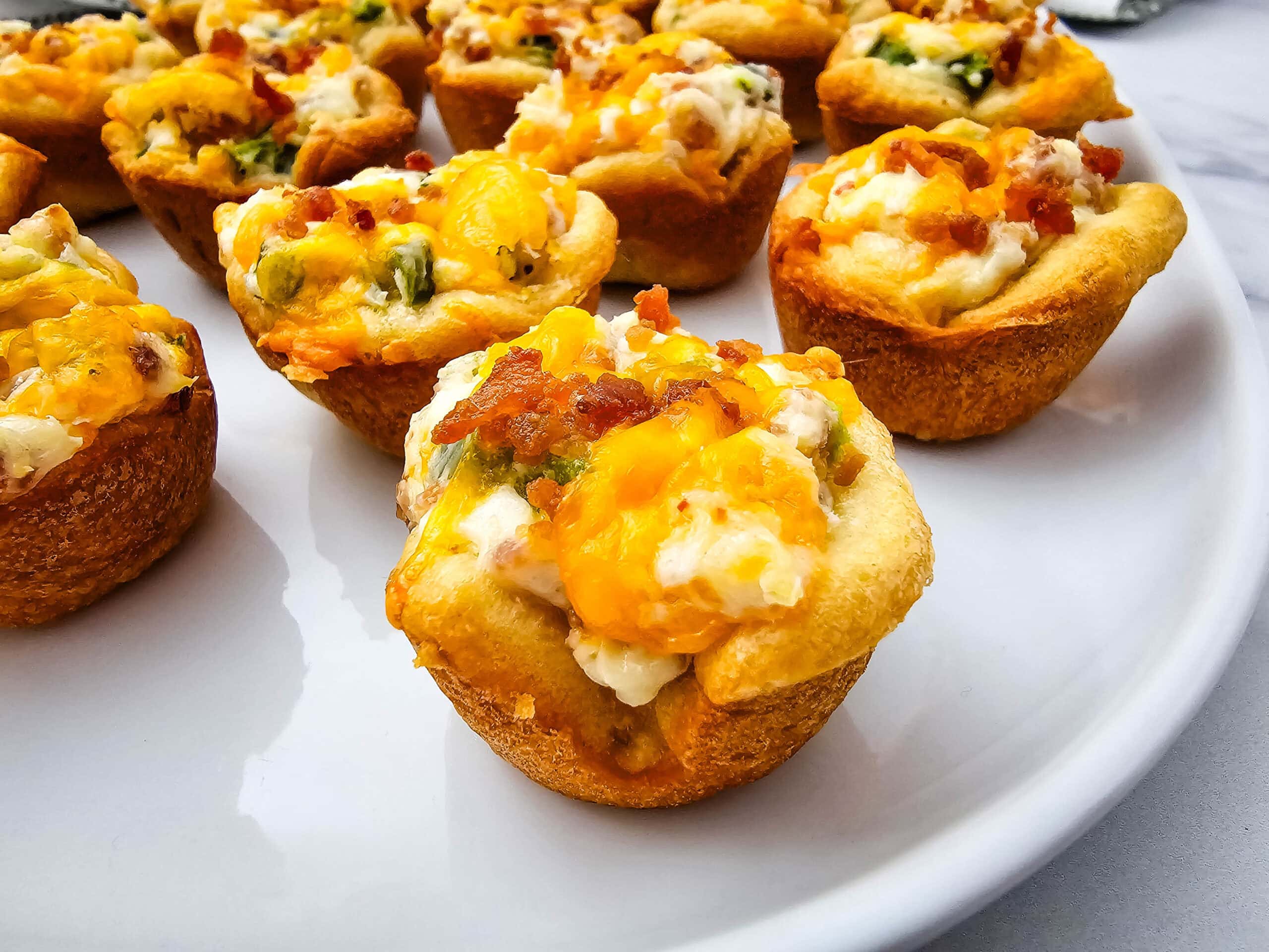 A close-up of savory baked muffin cups filled with creamy cheese, chopped green onions, and bacon bits, arranged on a white plate.