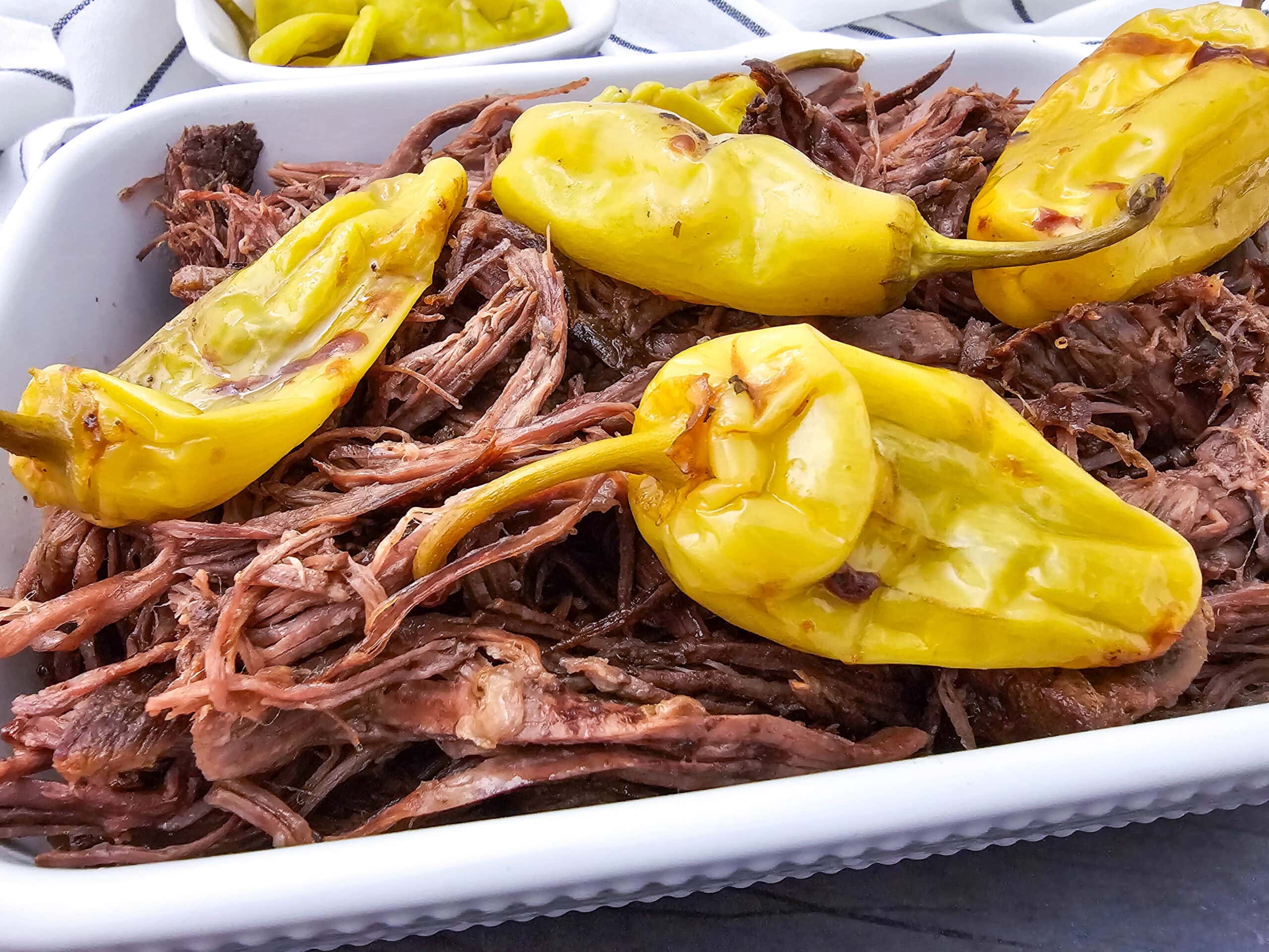 Shredded beef roast topped with several whole yellow pepperoncini peppers served in a white dish.