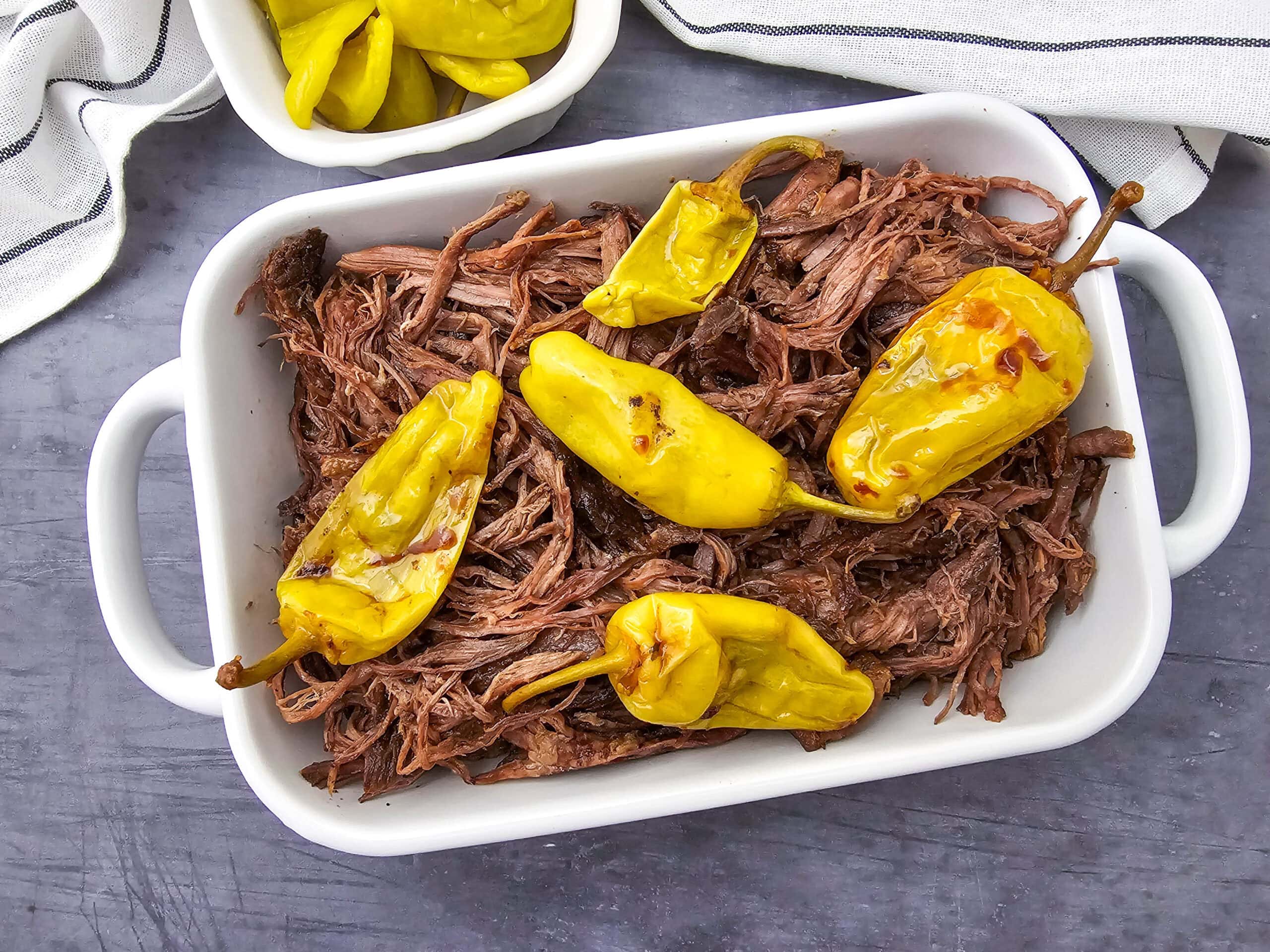 Shredded beef topped with whole yellow pepperoncini peppers served in a white rectangular baking dish, with a bowl of extra pepperoncinis and a striped cloth nearby on a gray surface.