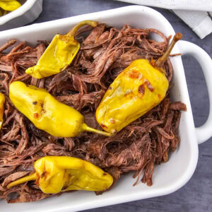 Shredded beef topped with whole yellow pepperoncini peppers in a white baking dish, placed on a dark surface with a striped cloth and a partial dish of more peppers nearby.