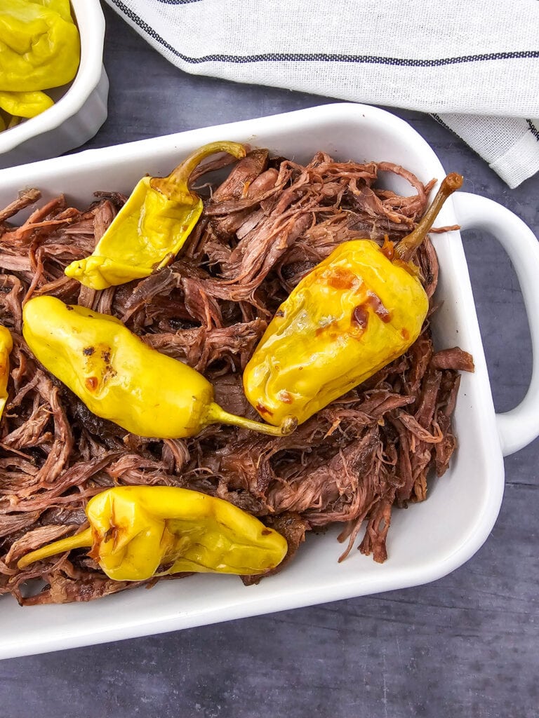 Shredded beef topped with whole yellow pepperoncini peppers in a white baking dish, placed on a dark surface with a striped cloth and a partial dish of more peppers nearby.