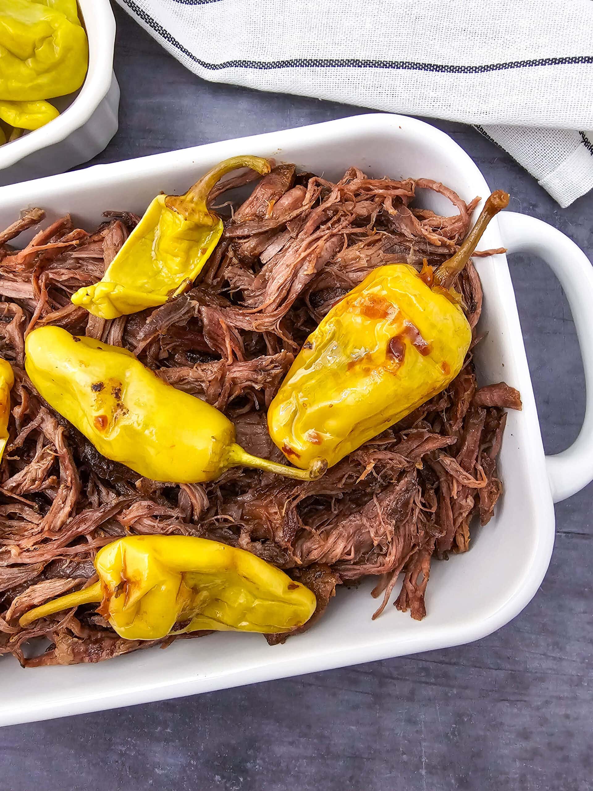 Shredded beef topped with whole yellow pepperoncini peppers in a white baking dish, placed on a dark surface with a striped cloth and a partial dish of more peppers nearby.