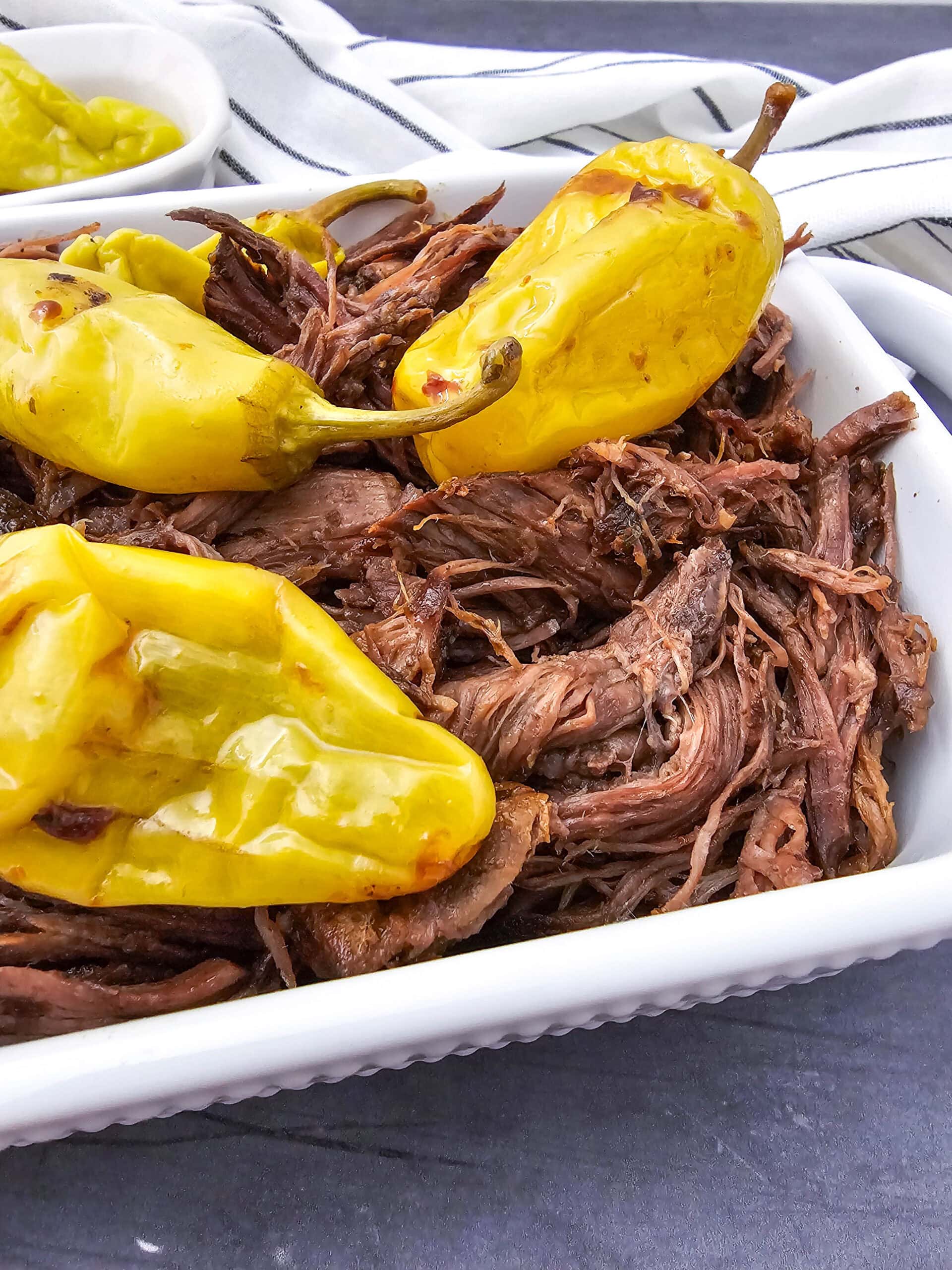 Shredded beef topped with whole yellow pepperoncini peppers in a white dish, with a striped cloth and a small bowl of sliced peppers in the background.