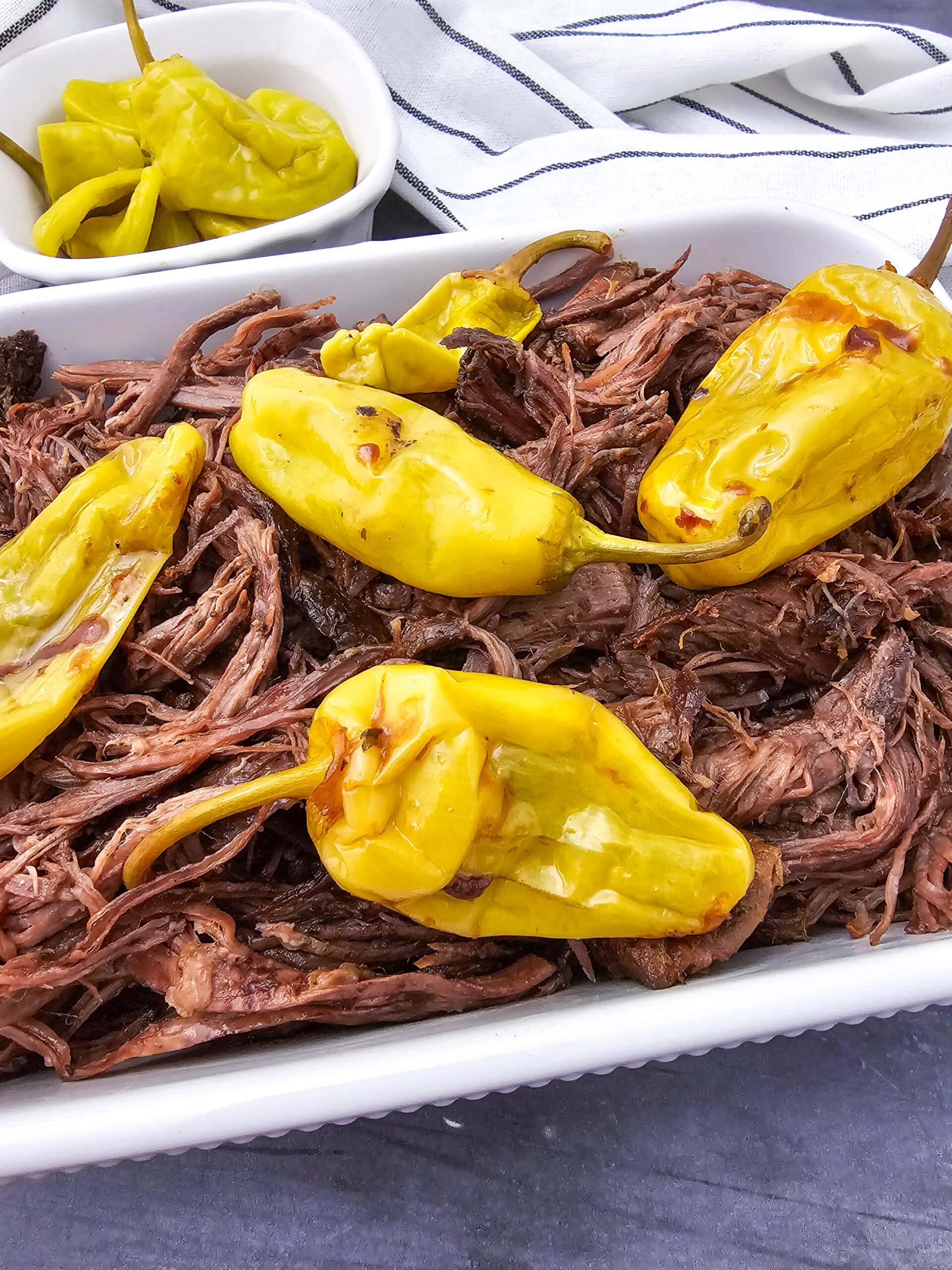 Shredded beef roast topped with whole yellow pepperoncini peppers in a white dish, with a small bowl of extra pepperoncinis in the background on a striped cloth.