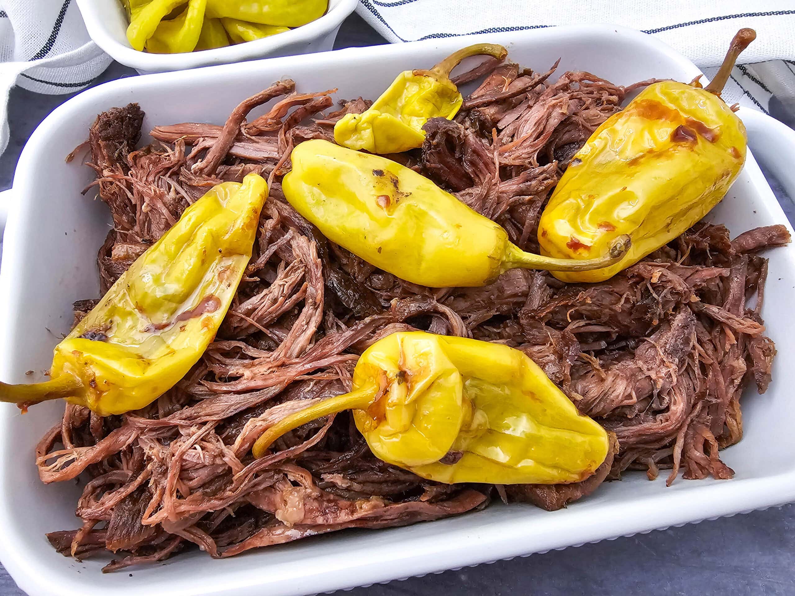 Shredded beef topped with whole yellow pepperoncini peppers in a white baking dish. A small portion of additional pepperoncini peppers is visible in a dish in the background.