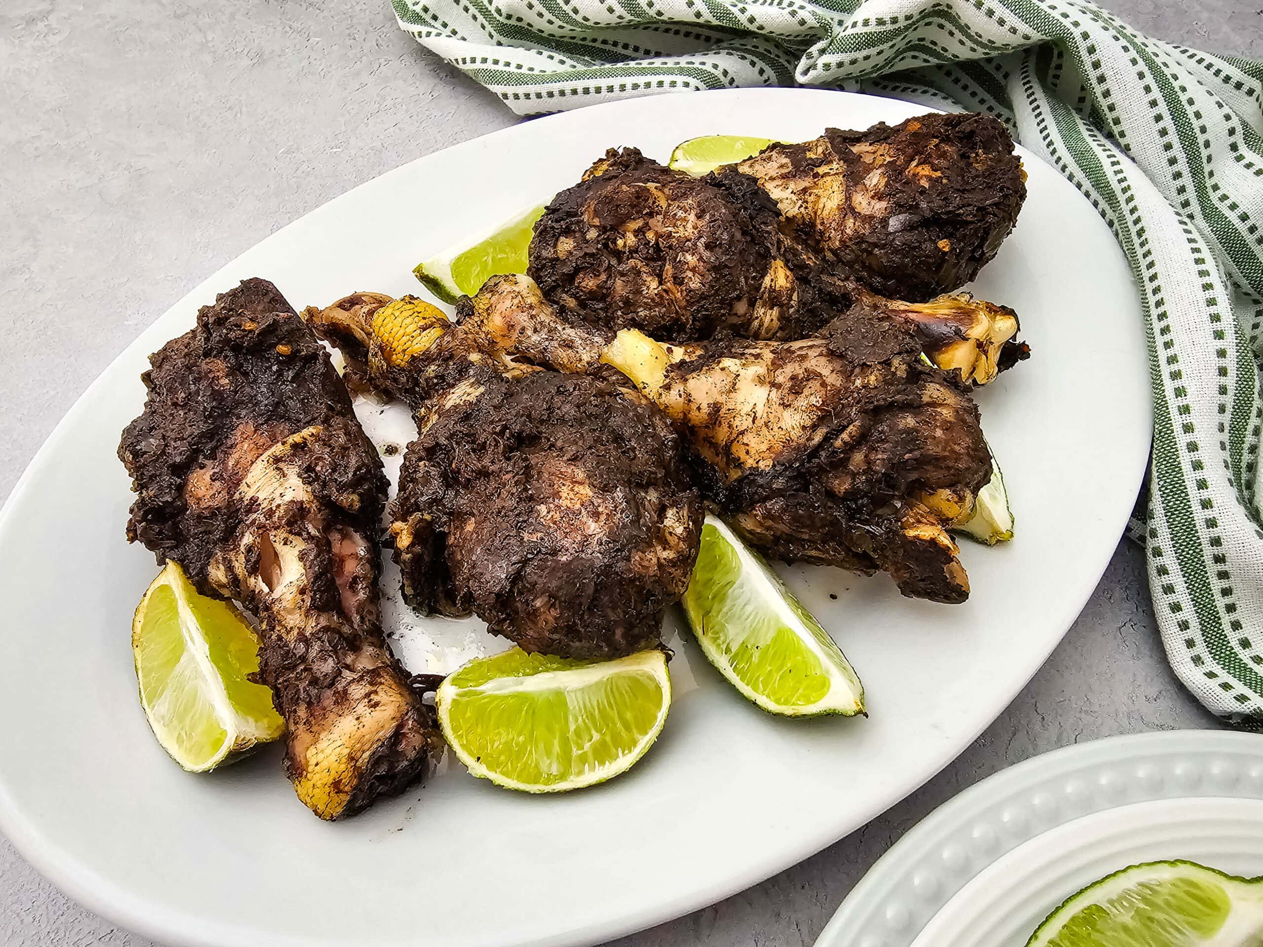 Four pieces of darkly seasoned grilled chicken are arranged on a white oval plate with lime wedges, set on a light surface with a green and white striped cloth in the background.