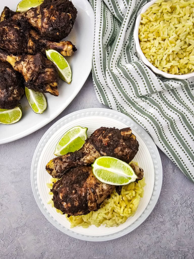 A plate with yellow rice topped with two pieces of seasoned grilled chicken and lime wedges, next to a bowl of rice and a platter of more chicken, all on a gray surface with a striped cloth.