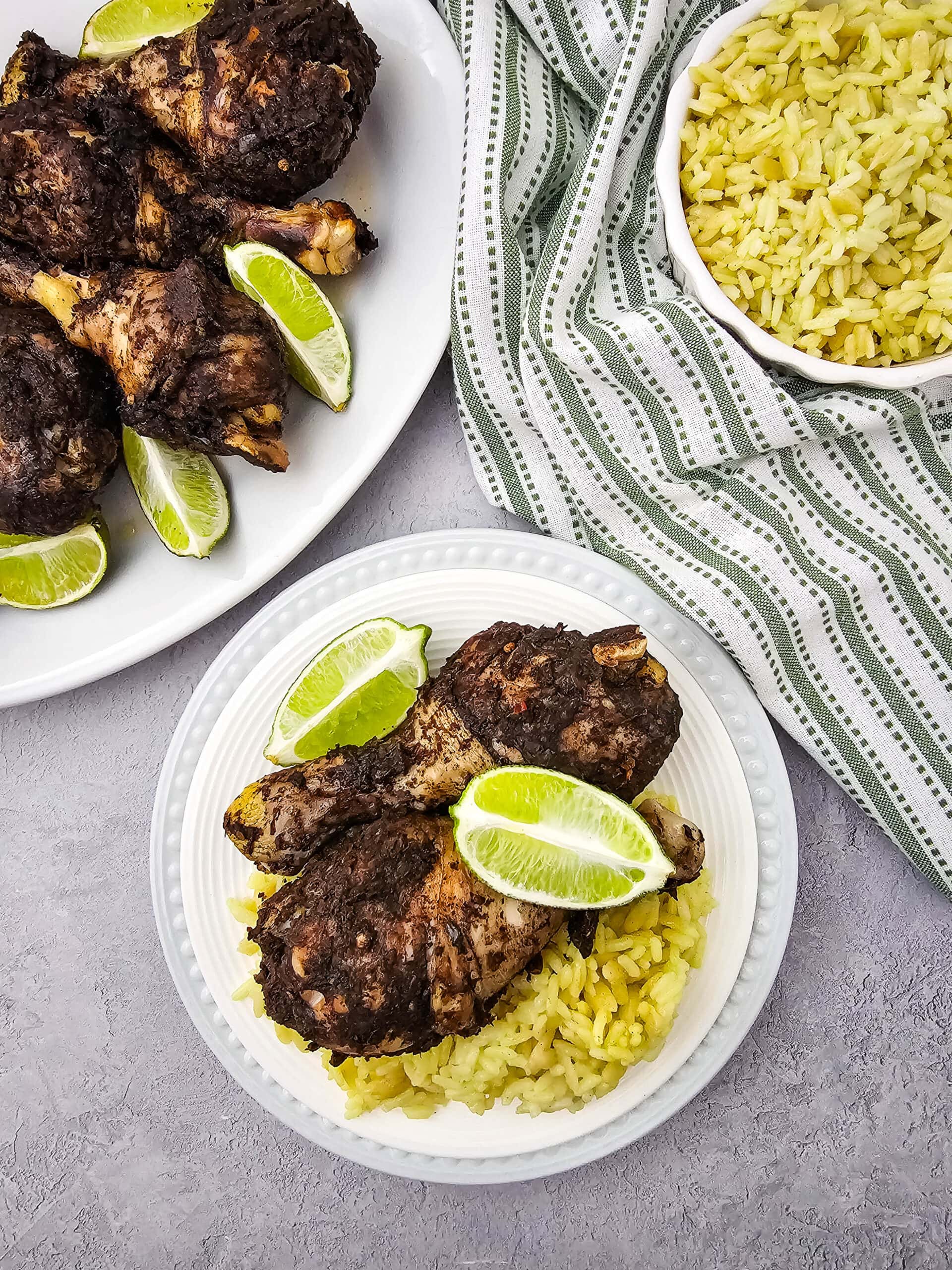 A plate with yellow rice topped with two pieces of seasoned grilled chicken and lime wedges, next to a bowl of rice and a platter of more chicken, all on a gray surface with a striped cloth.