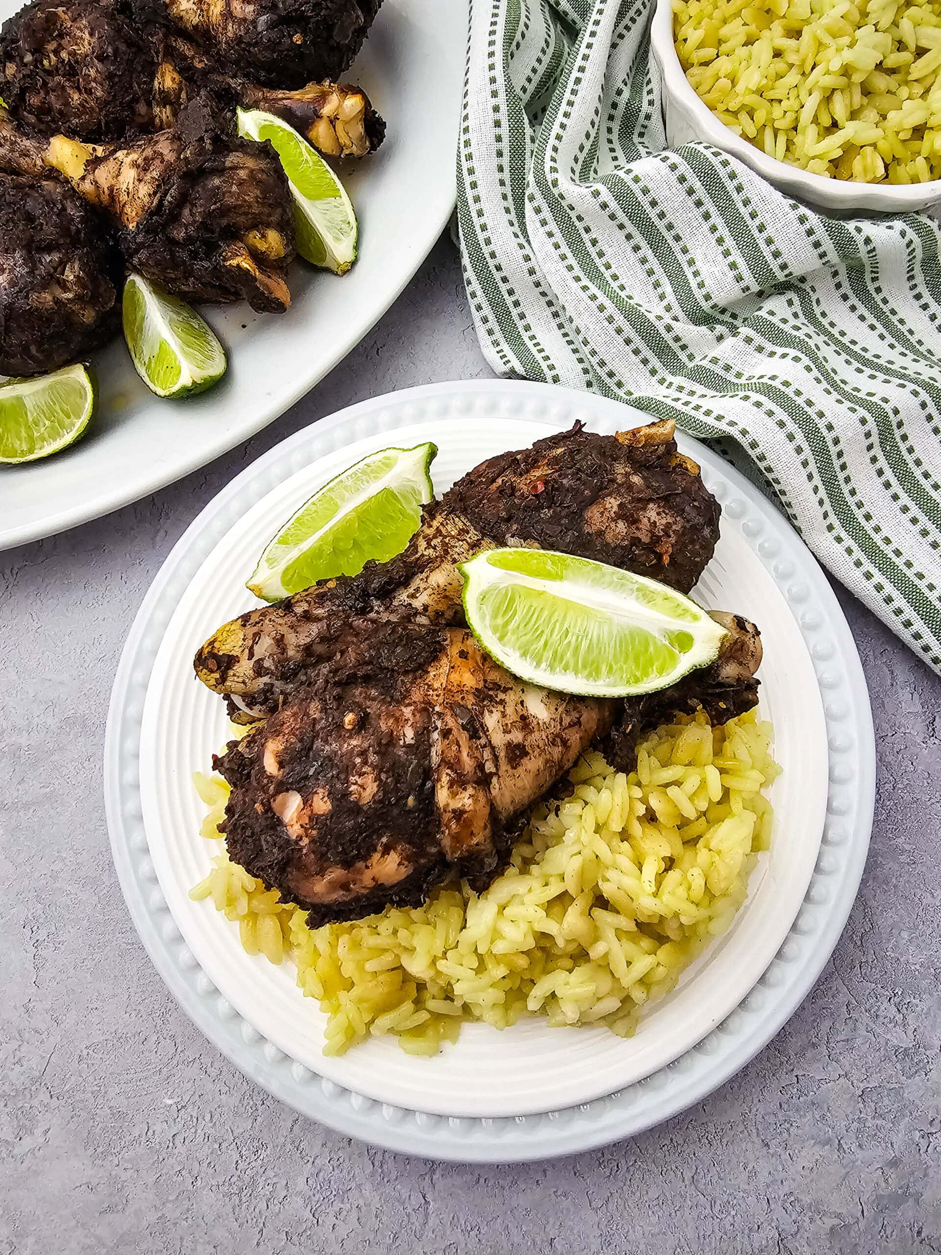 A white plate with two pieces of seasoned chicken atop yellow rice, garnished with lime wedges. A striped cloth, a bowl of rice, and another plate of chicken with lime wedges are in the background.
