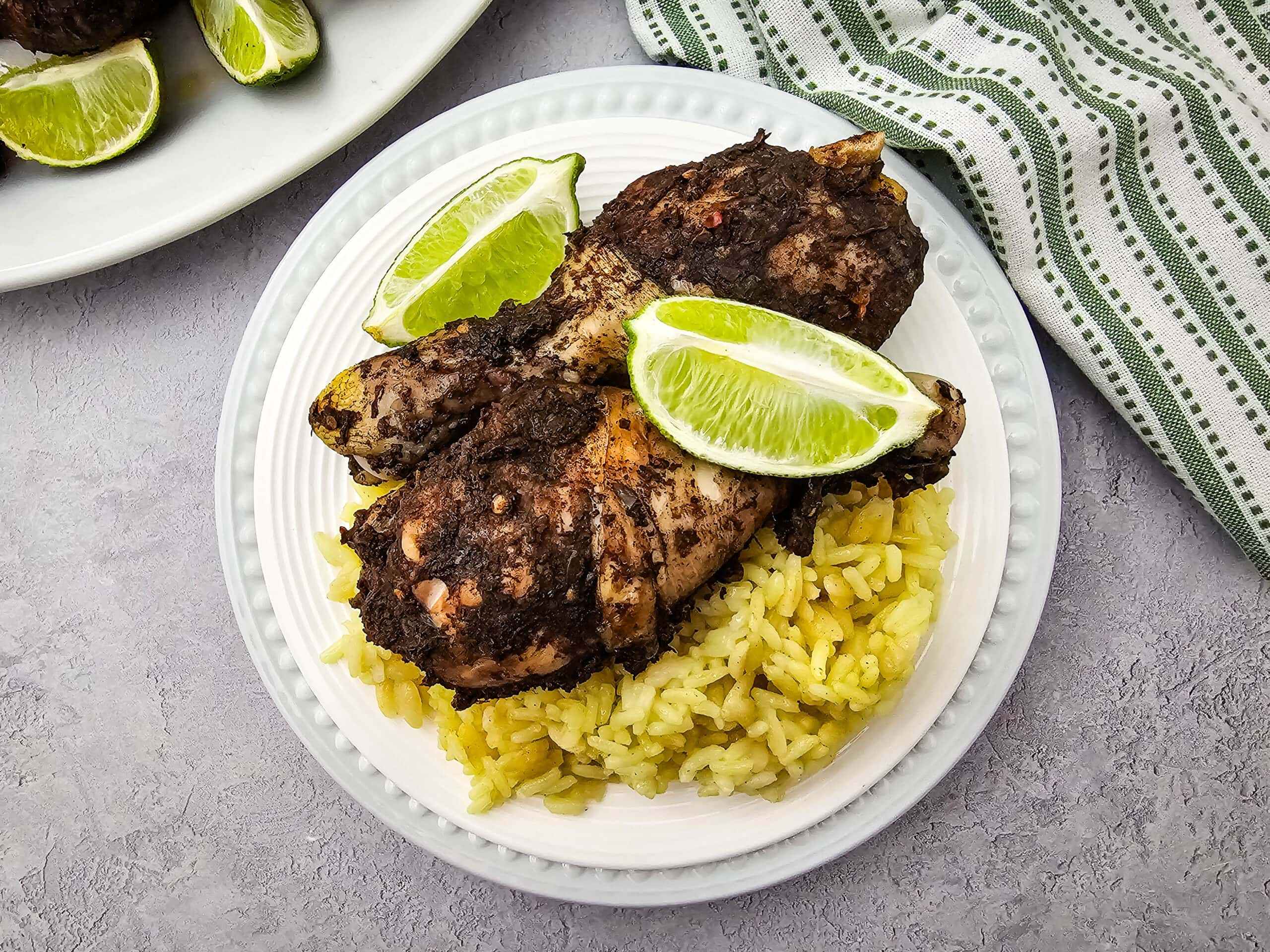 A white plate with yellow rice topped with two pieces of darkly seasoned chicken and two lime wedges, set on a gray surface next to a green-striped towel.