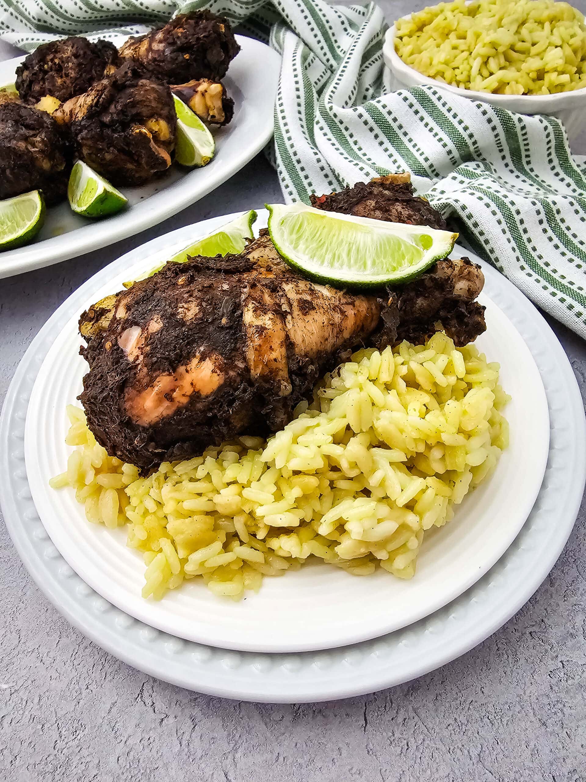 A plate of seasoned chicken drumstick topped with a lime wedge, served over a bed of yellow rice. In the background, there are more chicken drumsticks, a bowl of rice, and a green-striped cloth.