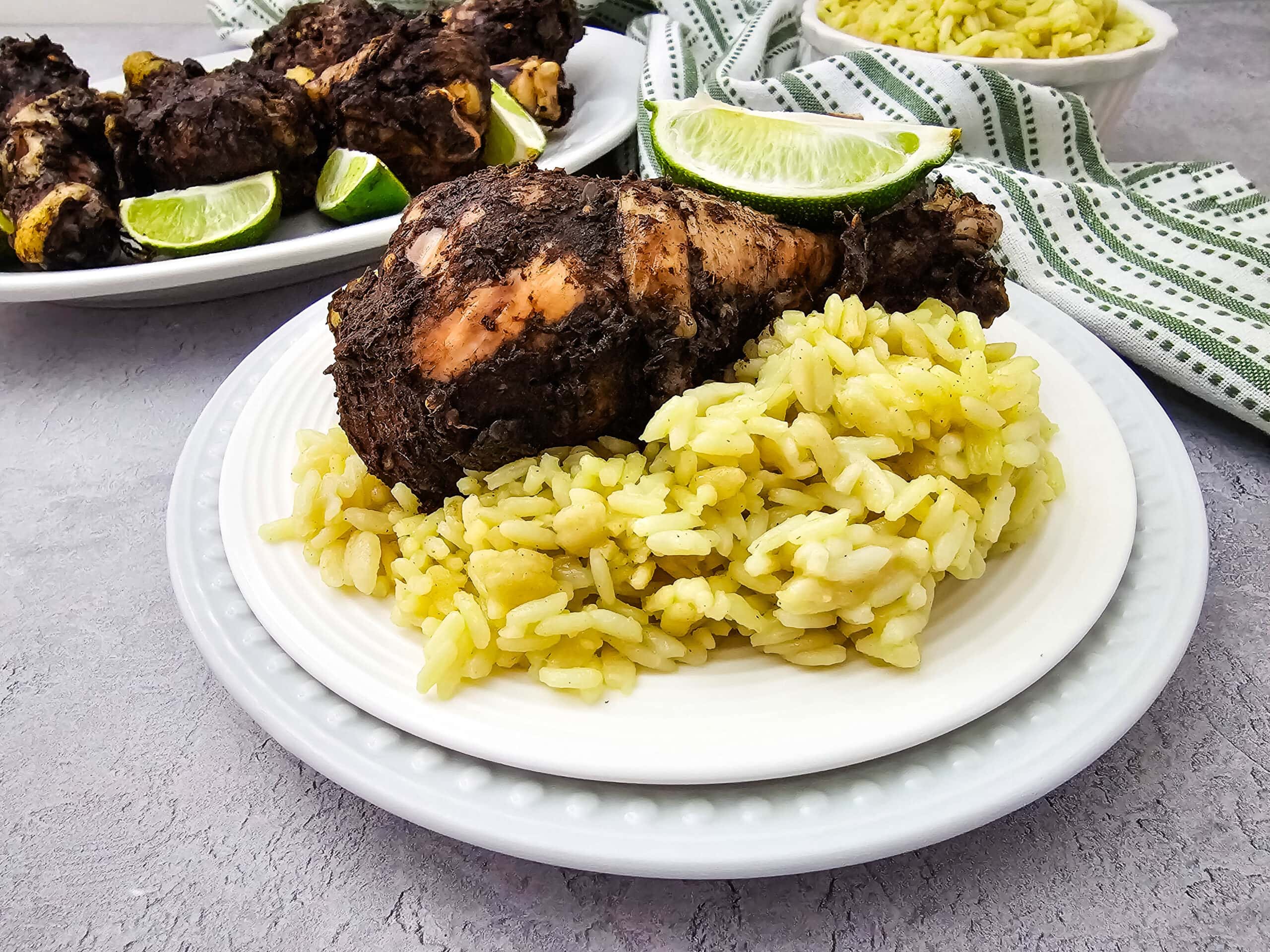 A plate with a serving of yellow rice and a piece of roasted chicken seasoned with dark spices, garnished with lime wedges. More chicken, lime, and rice are in the background on a plate and in a bowl.