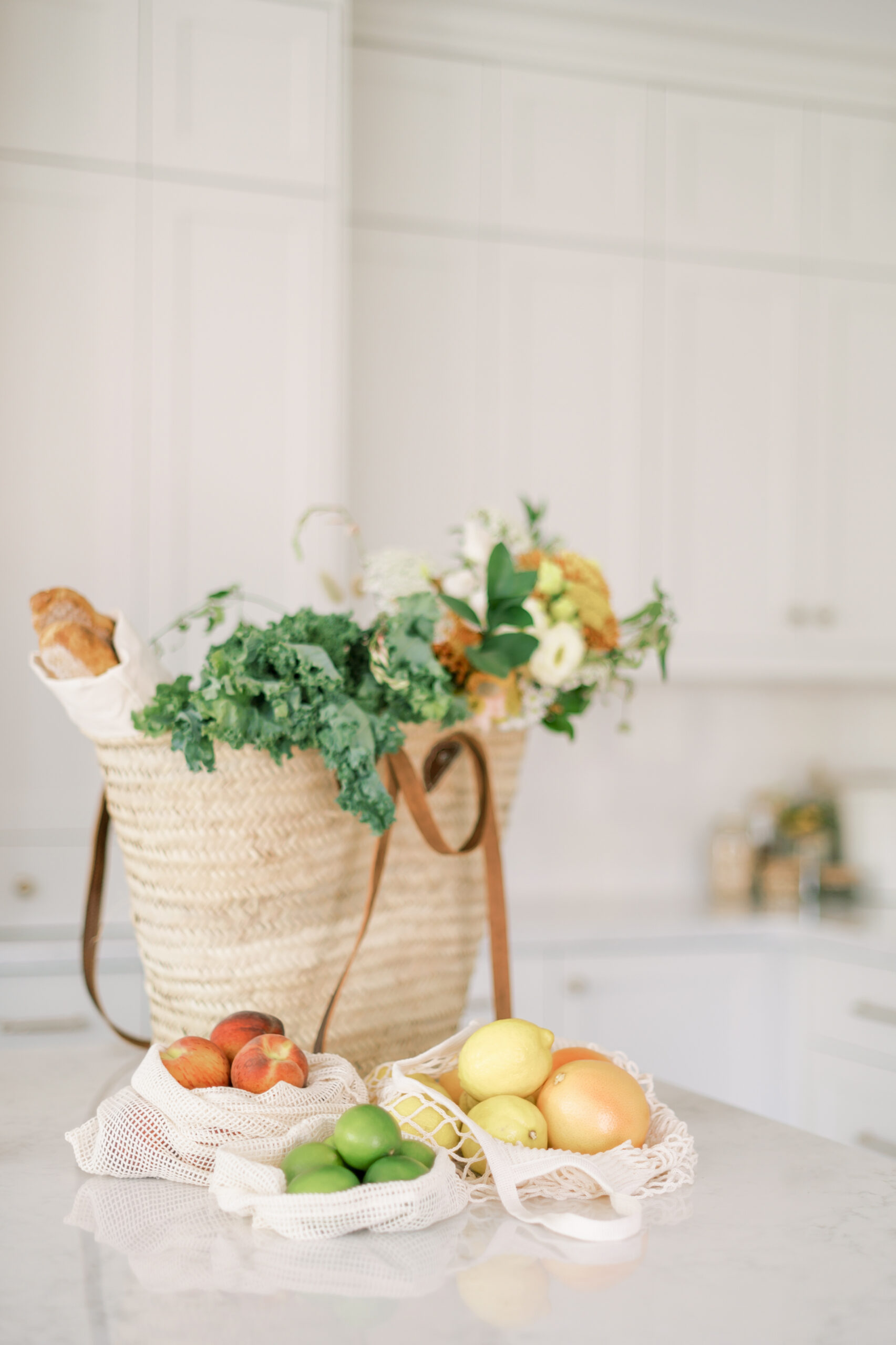 A woven market bag filled with flowers, greens, and a baguette sits on a light kitchen counter. Mesh bags with peaches, green apples, lemons, and an orange rest in front; white cabinets brighten the background.