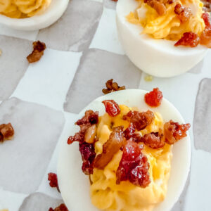 Close-up of deviled eggs topped with crispy bacon bits on a white and gray checkered surface. The eggs are halved, filled with a creamy yellow yolk mixture, and garnished with small bacon pieces.