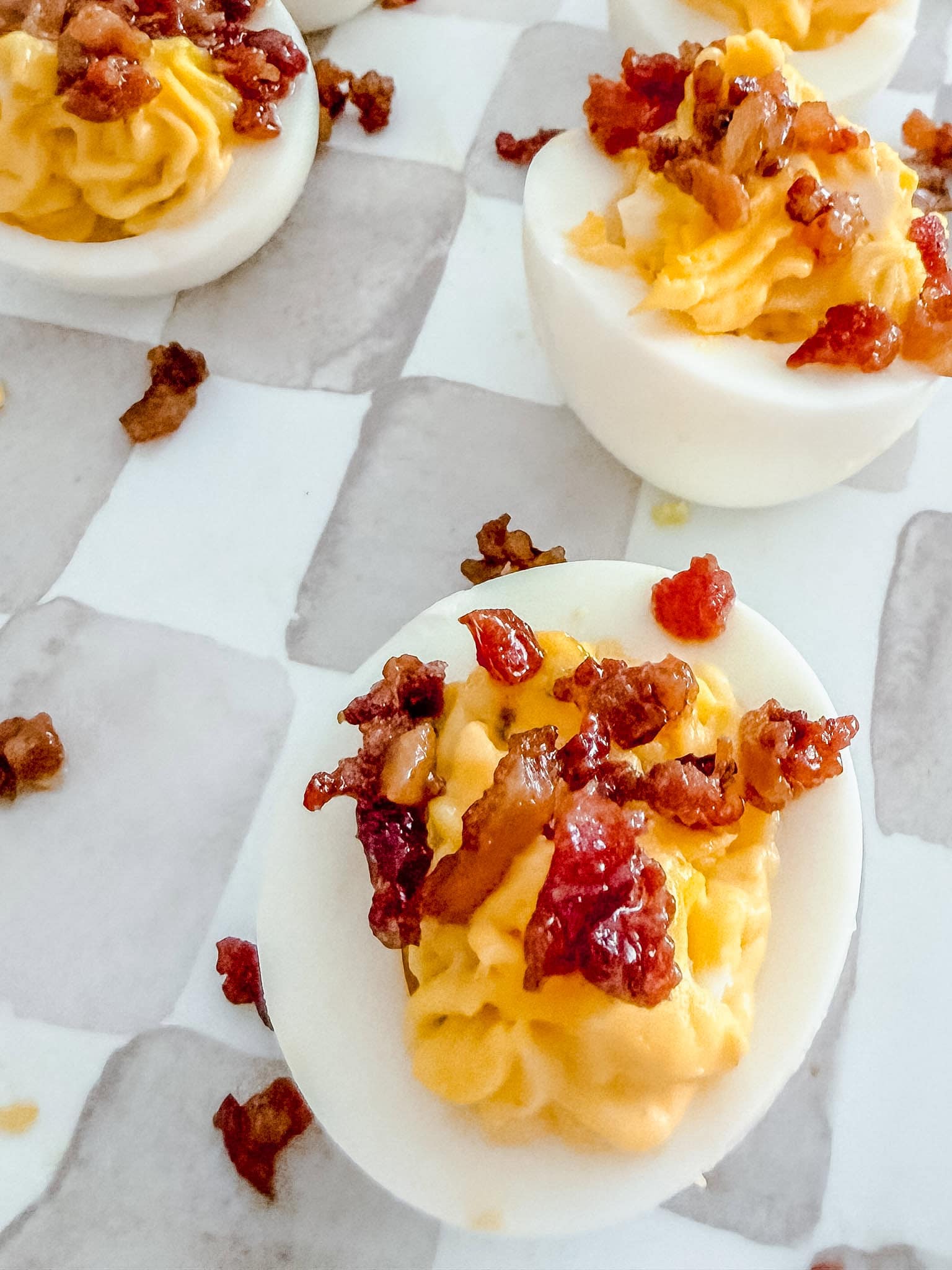 Close-up of deviled eggs topped with crispy bacon bits on a white and gray checkered surface. The eggs are halved, filled with a creamy yellow yolk mixture, and garnished with small bacon pieces.