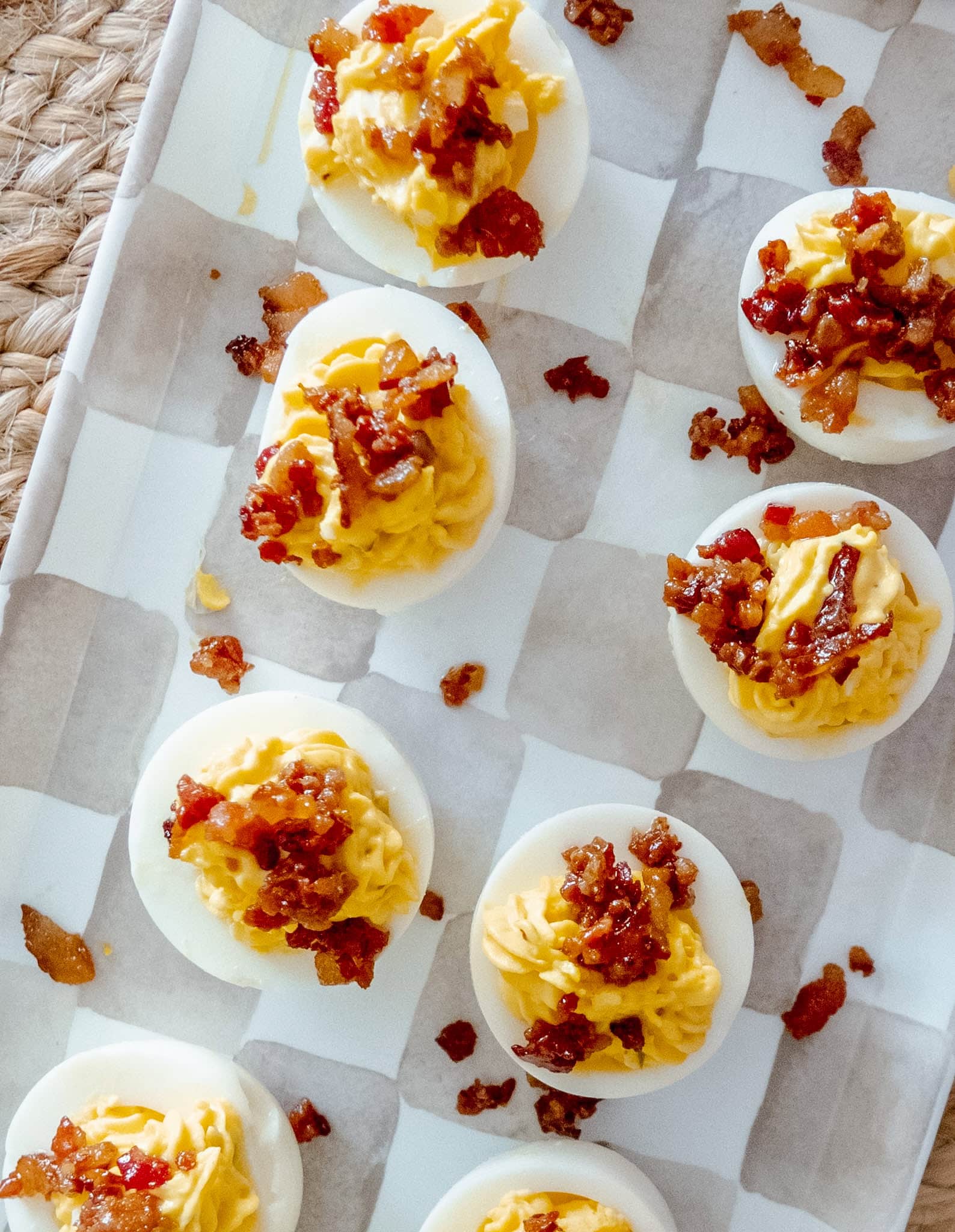 Deviled eggs topped with crispy bacon bits are arranged in rows on a white tray lined with parchment paper. The eggs have creamy yellow filling and a crunchy bacon garnish.