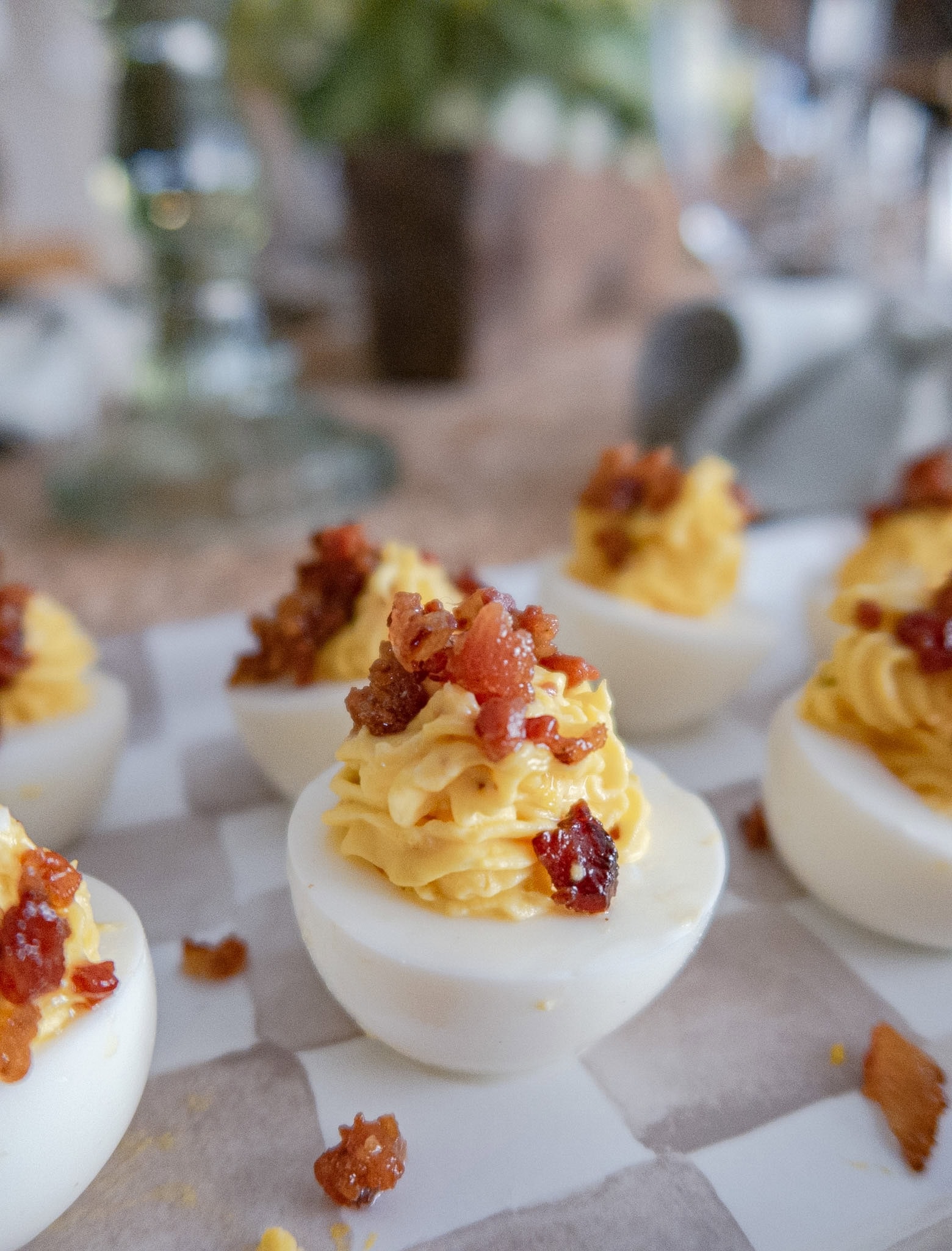 Close-up of deviled eggs topped with crispy bacon bits, arranged on a patterned platter. The eggs are neatly piped with a creamy yellow filling, and the background is softly blurred.