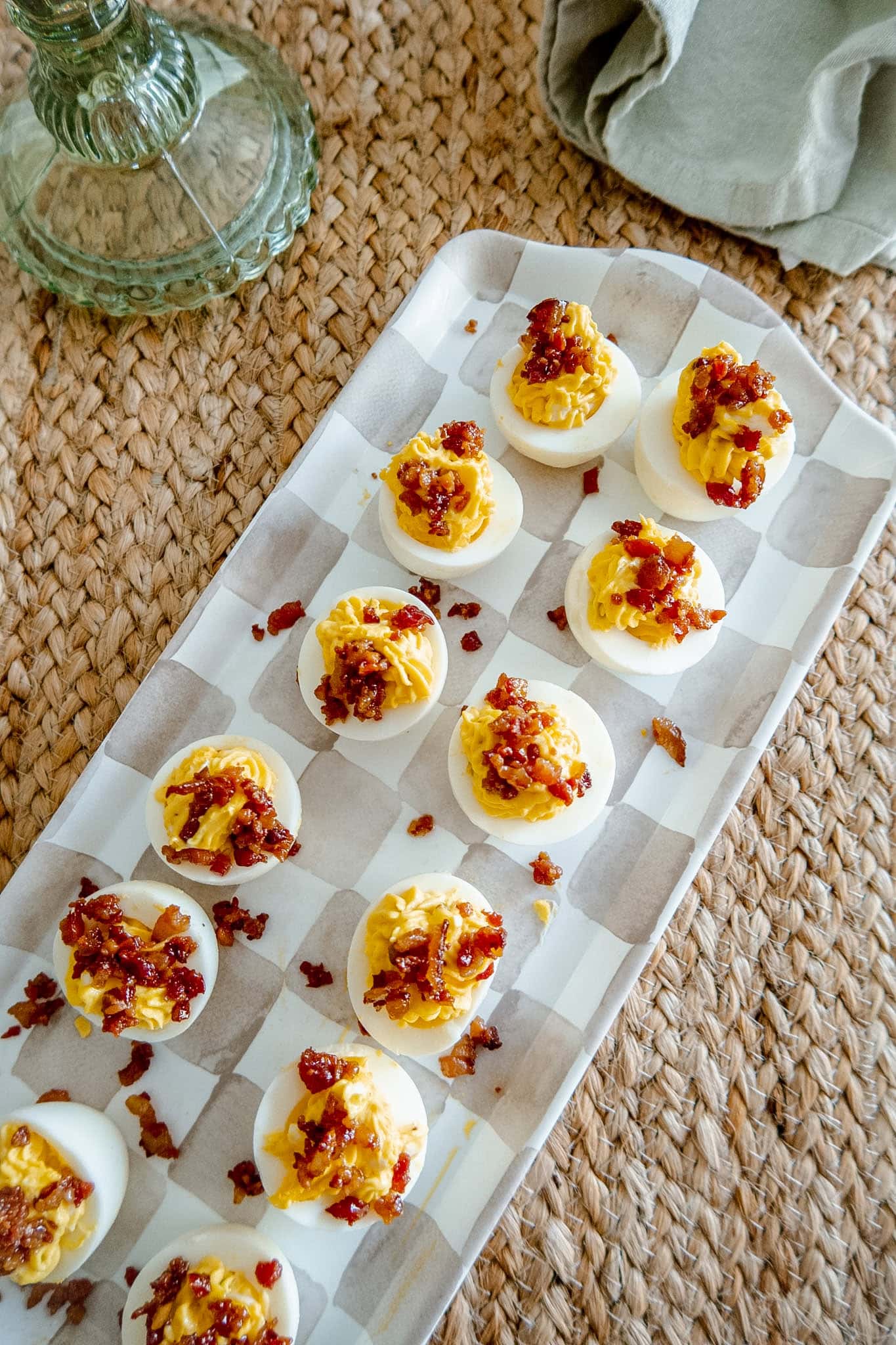A rectangular tray holds deviled eggs topped with crumbled bacon, arranged neatly on a checkered parchment paper. The tray sits on a woven placemat next to a green glass object and a folded beige napkin.