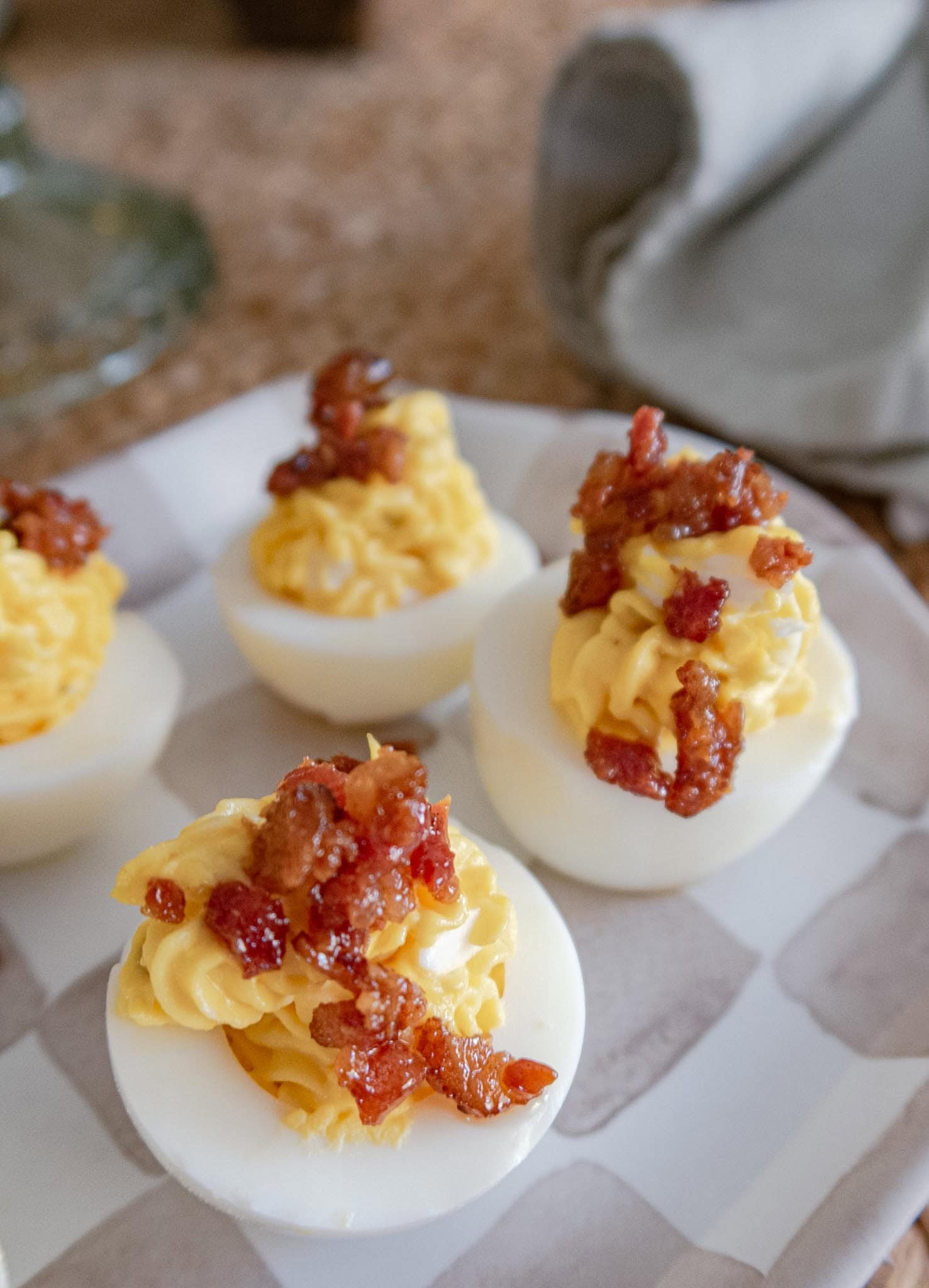 Four deviled eggs topped with crispy bacon bits are arranged on a white and light brown checkered plate, with a gray napkin in the background.