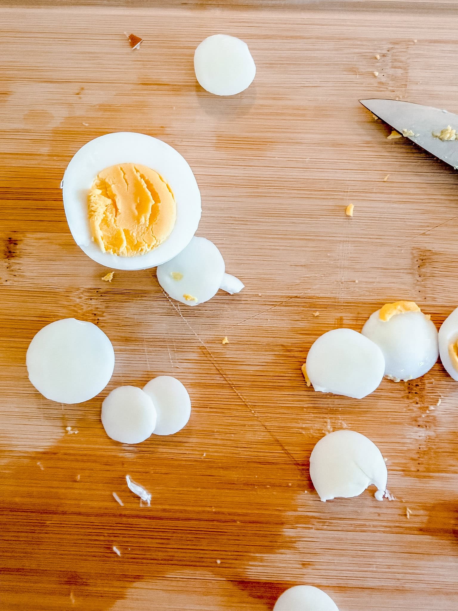 Sliced hard-boiled eggs on a wooden cutting board with a knife nearby; one egg is cut in half showing the yolk.