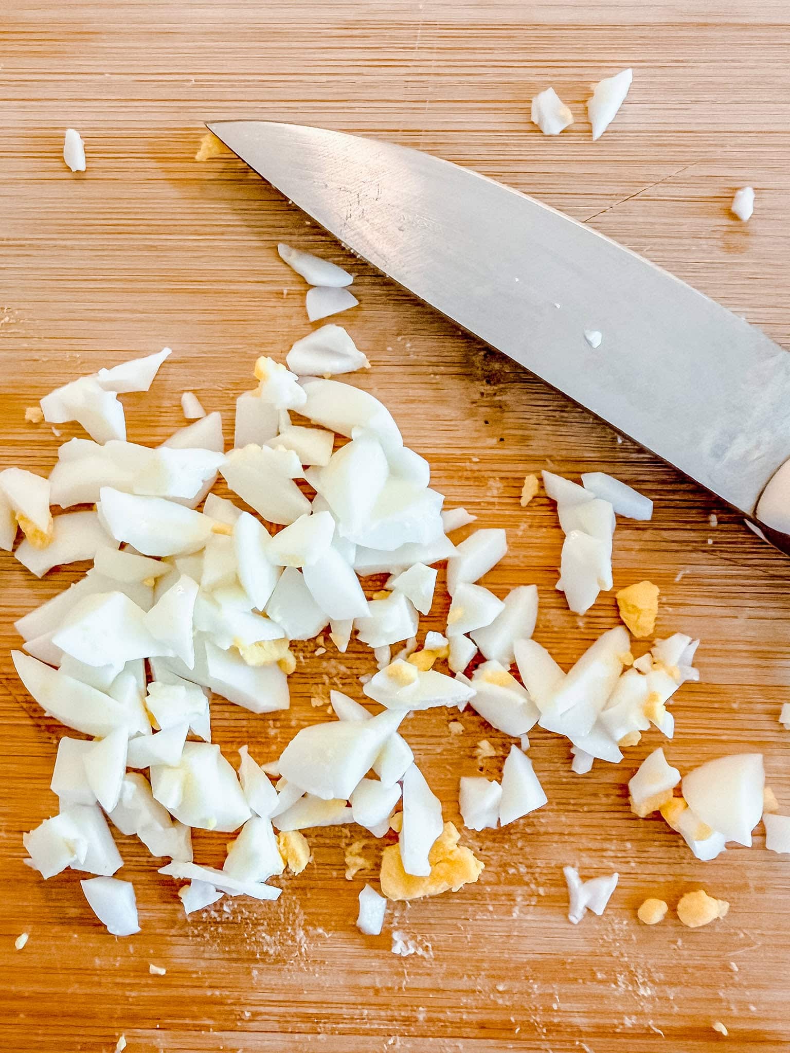 A chopped hard-boiled egg with pieces of white and yolk sits on a wooden cutting board next to a large kitchen knife.