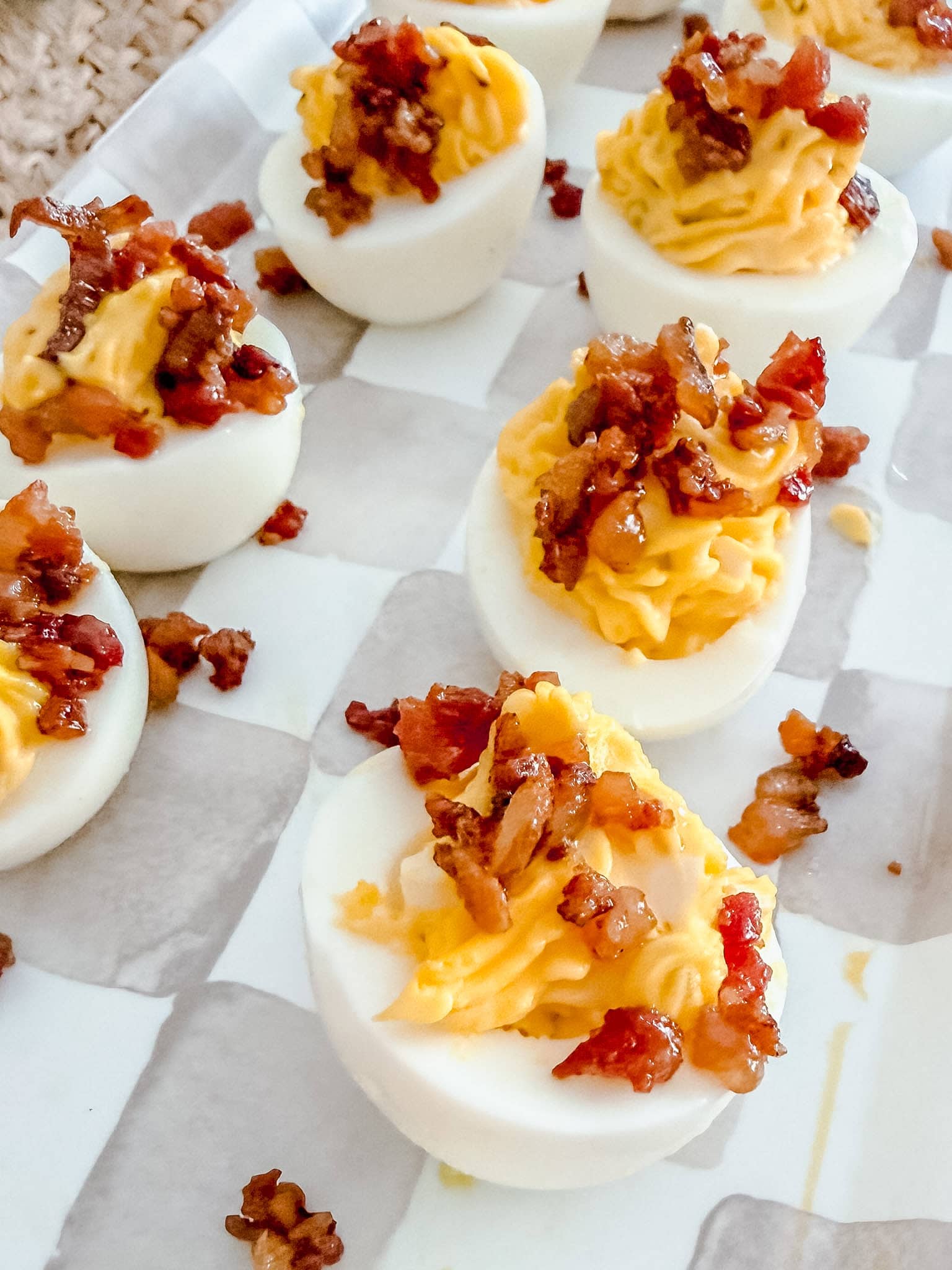 Deviled eggs topped with crispy bacon pieces are arranged on a white and gray checkered platter. The eggs have a creamy yellow filling and are garnished with bits of bacon.