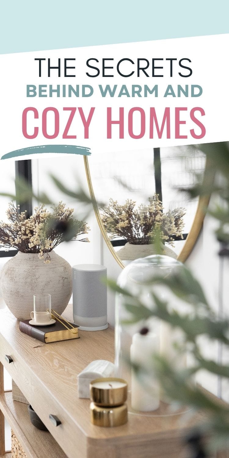 A wooden console table is decorated with vases of dried flowers, candles, and small decorative objects in front of a round mirror. Overlay text reads: The secrets behind warm and cozy homes.