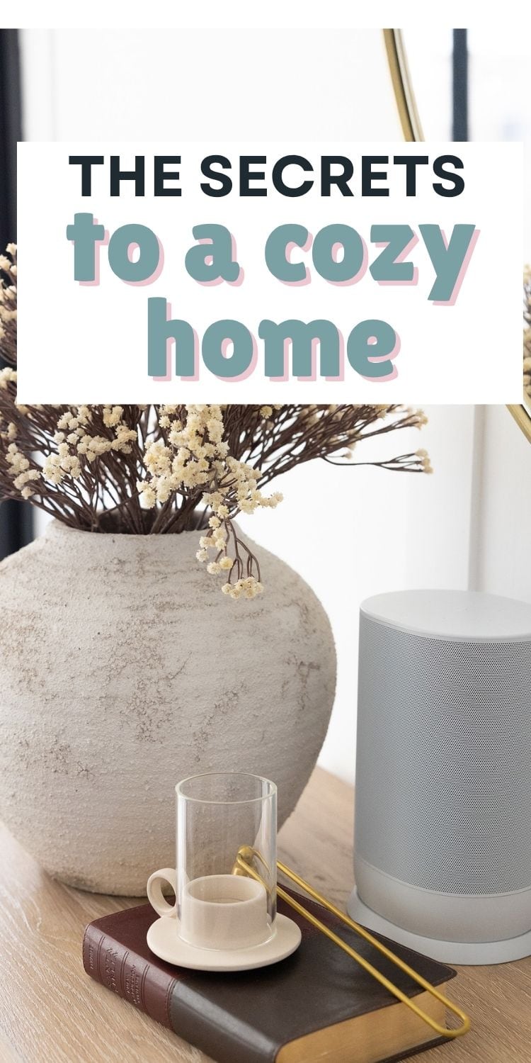 A beige vase with dried flowers, a white speaker, a glass cup and saucer with a tea bag, and two stacked books sit on a light wooden table. A sign above reads The secrets to a cozy home.