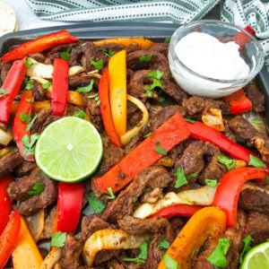 A sheet pan filled with cooked steak strips, red and yellow bell peppers, and onions, garnished with cilantro and lime slices. A small bowl of sour cream sits on the pan. A tortilla and a striped cloth are nearby.