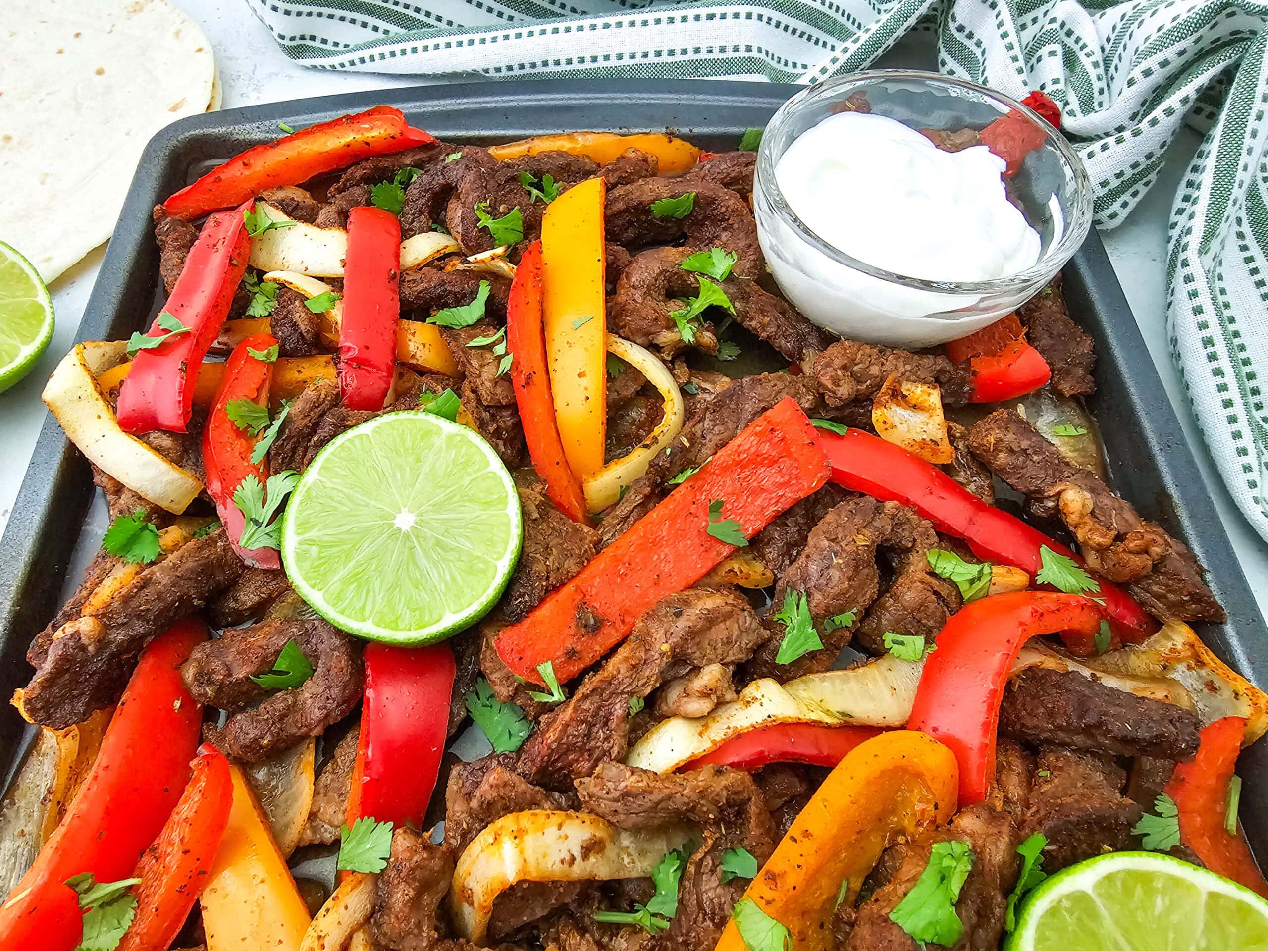 A sheet pan filled with cooked steak strips, red and yellow bell peppers, and onions, garnished with cilantro and lime slices. A small bowl of sour cream sits on the pan. A tortilla and a striped cloth are nearby.