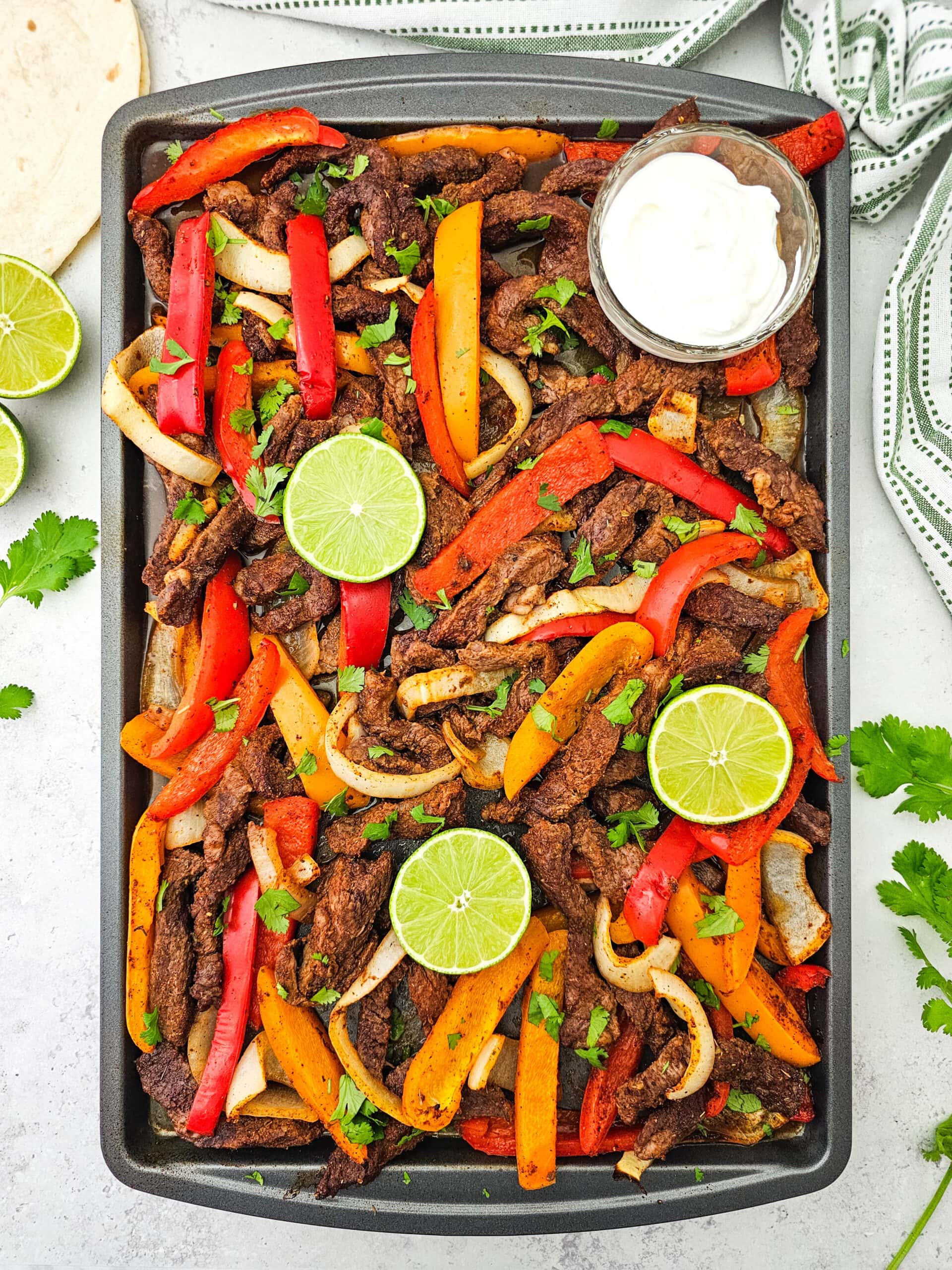 A sheet pan filled with cooked beef strips, red and yellow bell peppers, and onions, garnished with lime slices and cilantro. A small bowl of white sauce sits in one corner. Tortillas and lime halves are nearby.