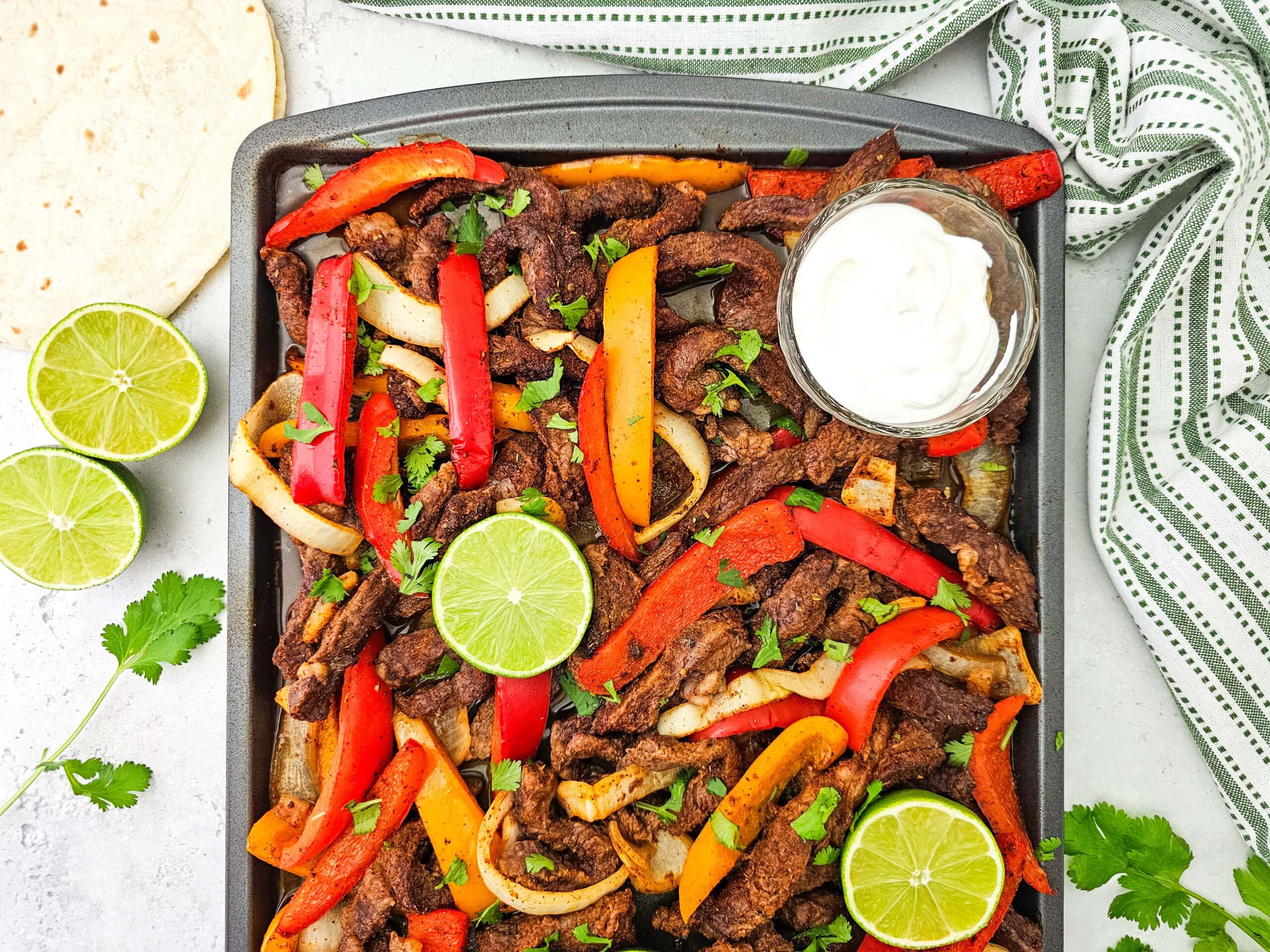 A sheet pan filled with beef fajitas, sliced red and yellow peppers, onions, and garnished with lime slices and cilantro. A small bowl of sour cream is on the pan, with lime halves and a tortilla on the side.