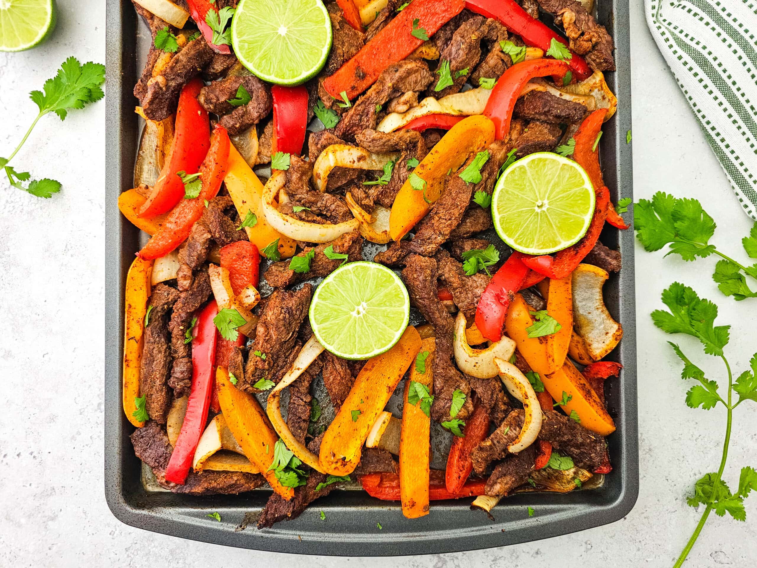 A tray of colorful beef fajitas with sliced red and yellow bell peppers, onions, topped with fresh cilantro and lime slices, set on a light surface with cilantro leaves and a striped towel nearby.
