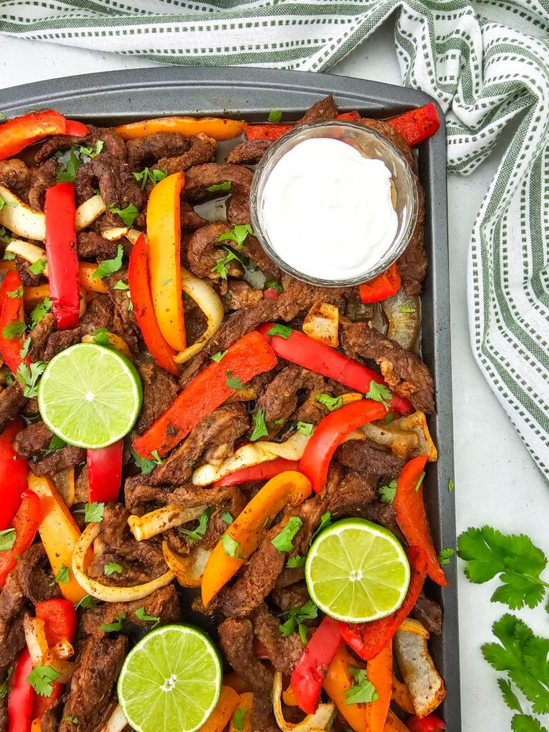 Sheet pan filled with sliced beef, red and yellow bell peppers, onions, garnished with fresh cilantro and lime slices. A small bowl of white sauce sits in one corner. A striped towel and cilantro leaves are nearby.