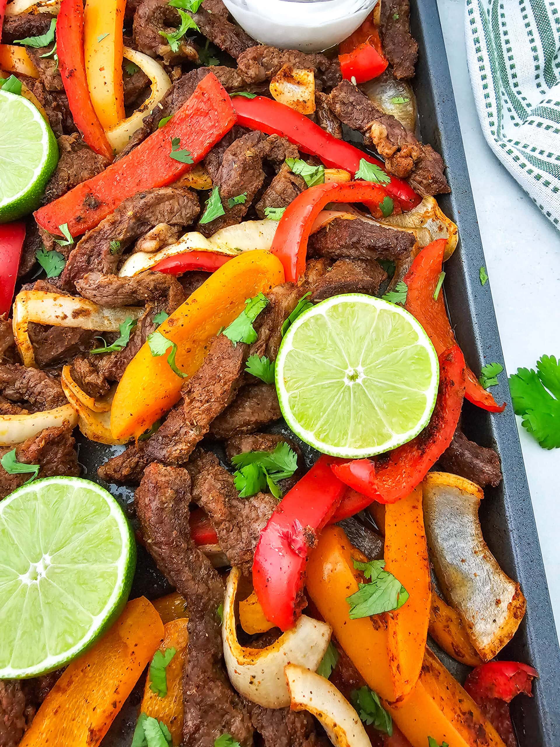 Colorful beef fajitas with sliced red and yellow bell peppers, onions, fresh cilantro, and lime slices on a baking tray. A bowl of white sauce and a striped cloth are visible in the background.