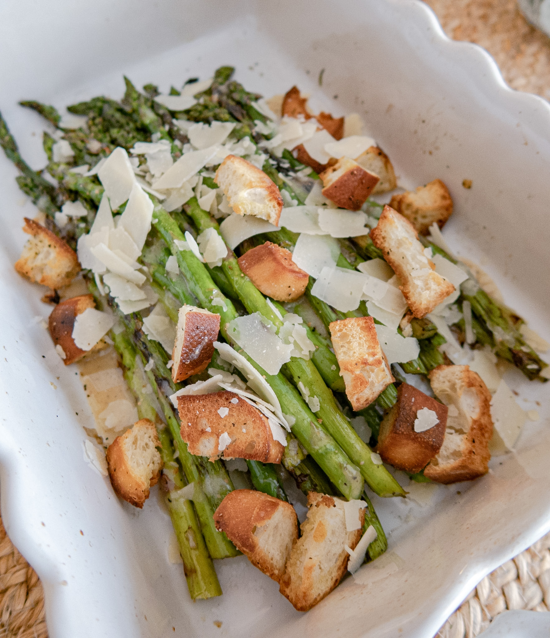Roasted asparagus spears topped with shaved parmesan cheese and toasted crouton pieces, served in a white ceramic baking dish.